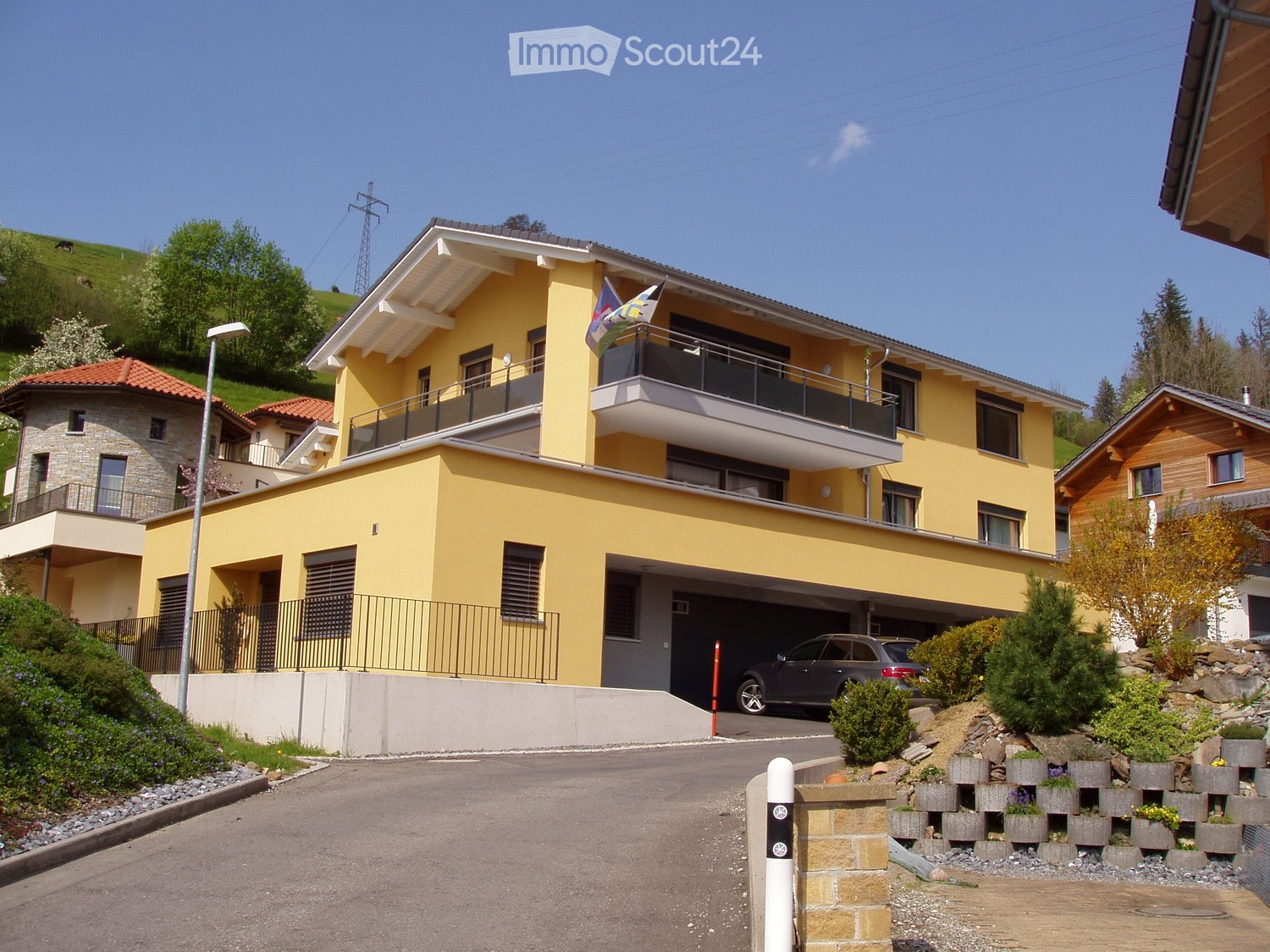Yellow two-story house, garage, metal fencing, plants, flag, car