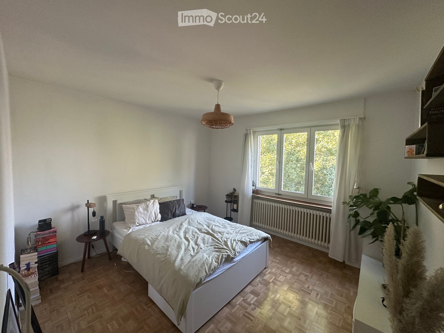 white walls, white bed with white bedding, white night table with lamp, white radiator, white shelf, wooden floor, window with white curtains