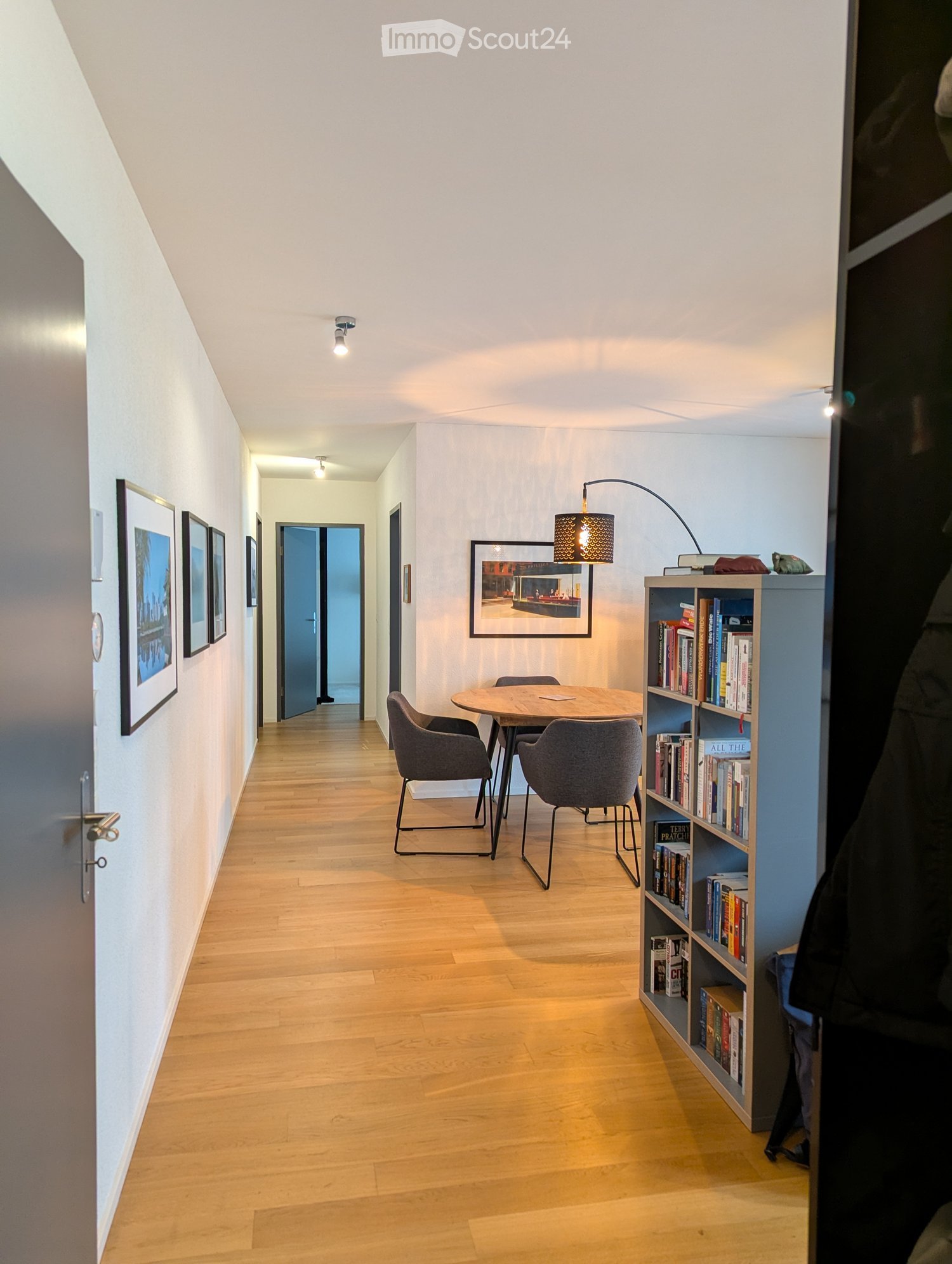 wooden floor, white walls, corridor, 2 picture frames, a wooden table, chairs, lamp, bookcase, bookshelves