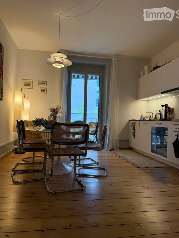 Dining area with chairs around a table, open kitchen with oven, floor lamp, and a window leading to balcony