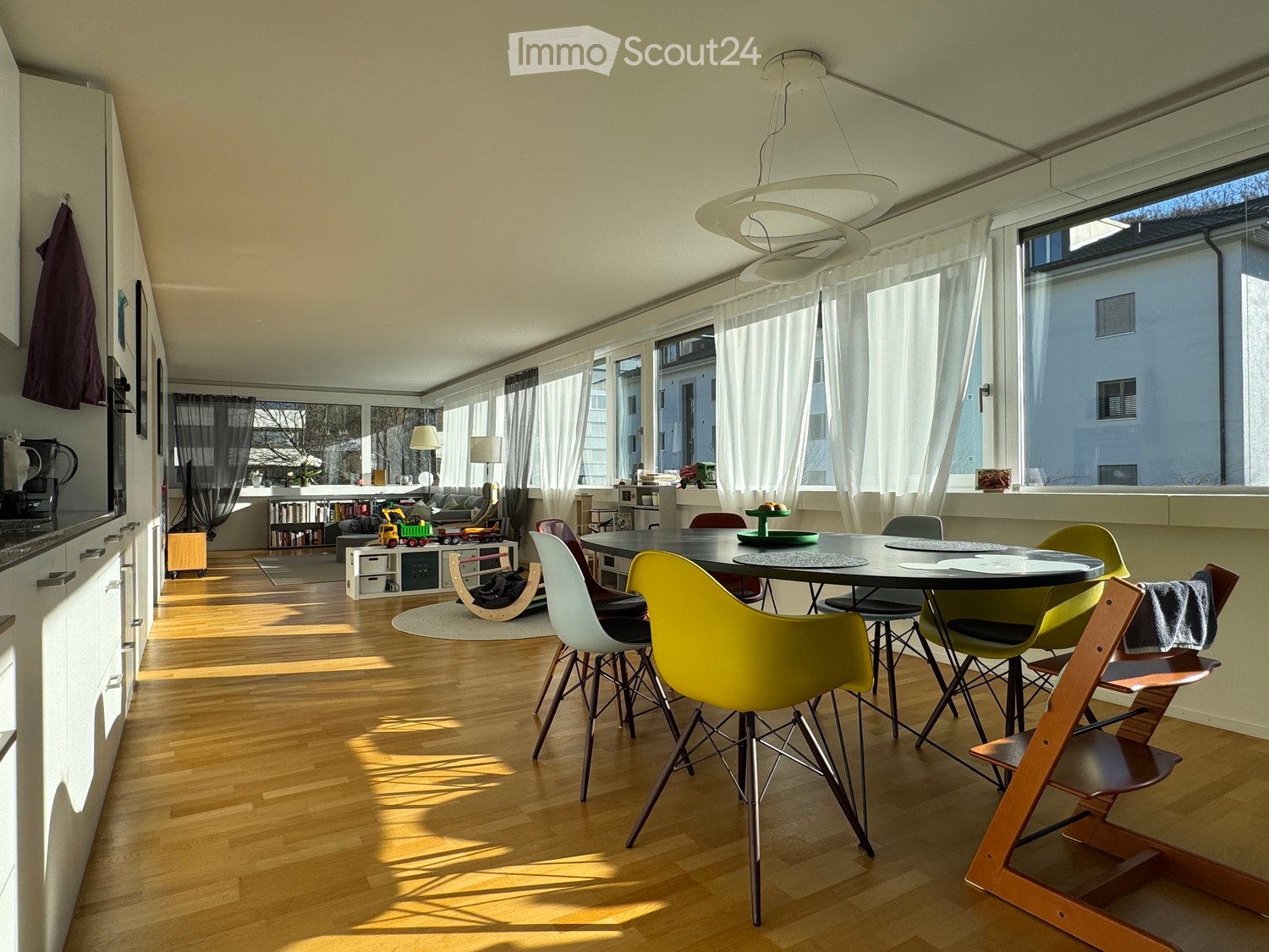 spacious room, wooden floor, white walls, modern furniture, dining table with colorful chairs, large windows with white curtains, kitchen area, bright sunlight