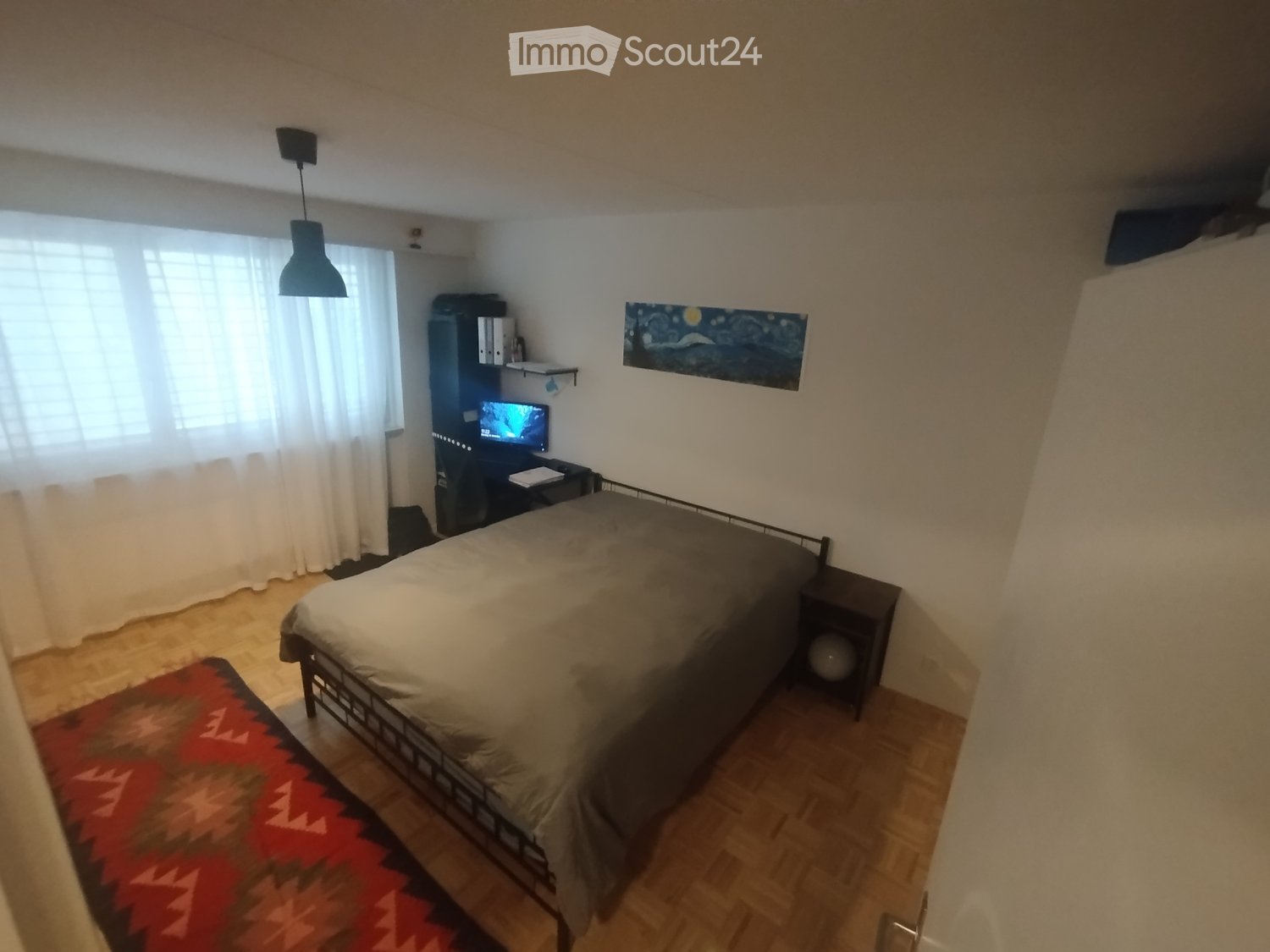 Bedroom: bed, rug, desk with monitor, nightstand, lamp, window with white curtains, painting on the wall