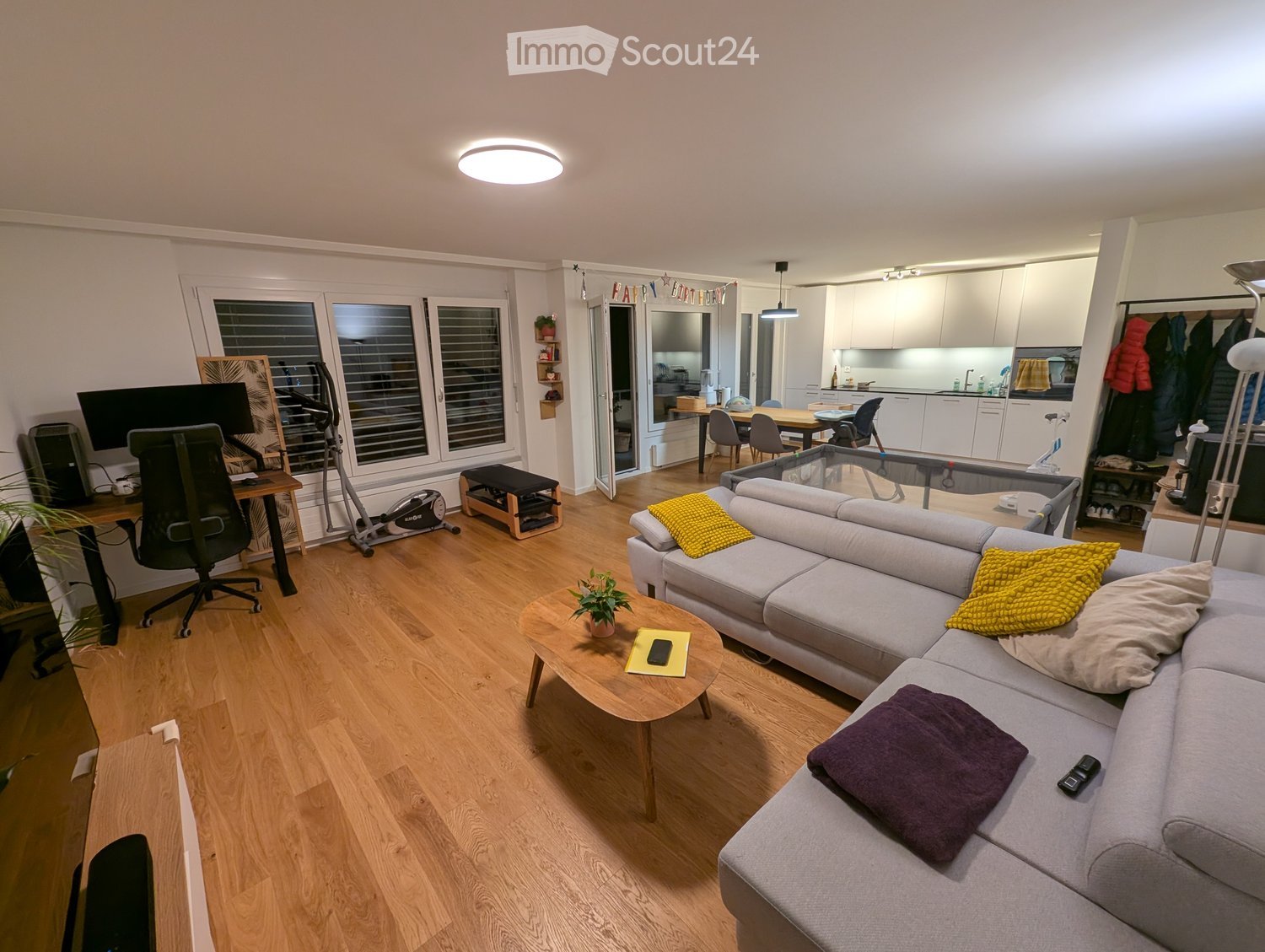 open living room with wooden floor, light grey sofa, round wooden coffee table with plant, desk with computer and chair, white cabinets, ceiling light