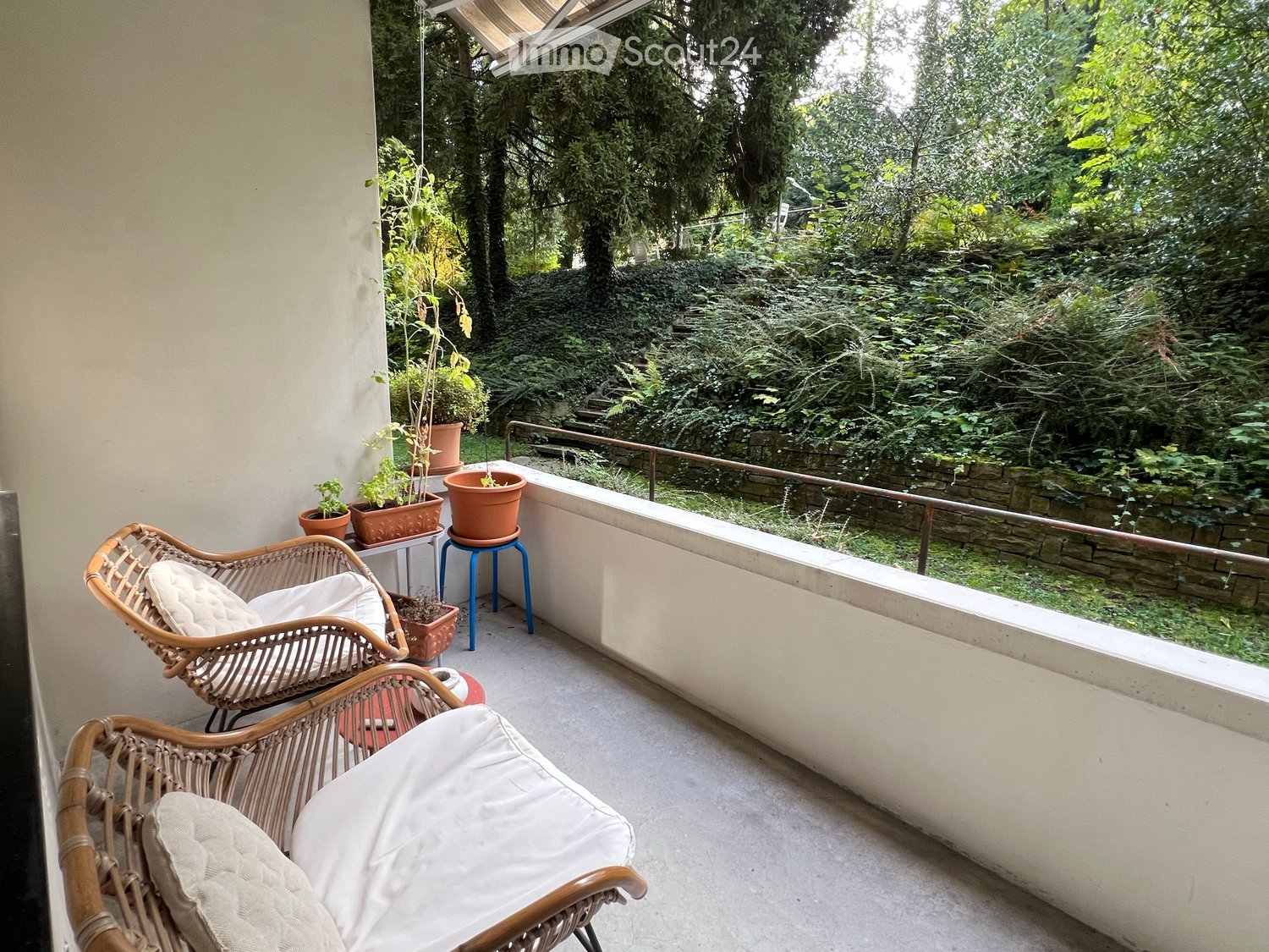 balcony with outdoor seating, small table, and potted plants, fenced area, green hill in the background