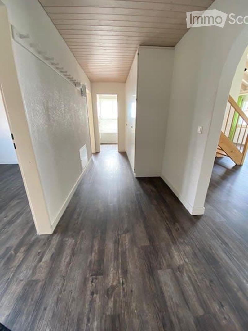The hallway has wooden floors, white walls, a wooden ceiling with visible beams, and a small window at the end of the hall. There is a wooden staircase with a green railing on the right side.