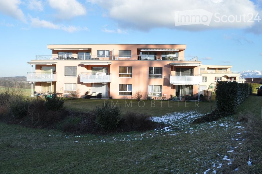 Multi-storey apartment building with balconies, surrounded by grass and bushes, a car parked nearby