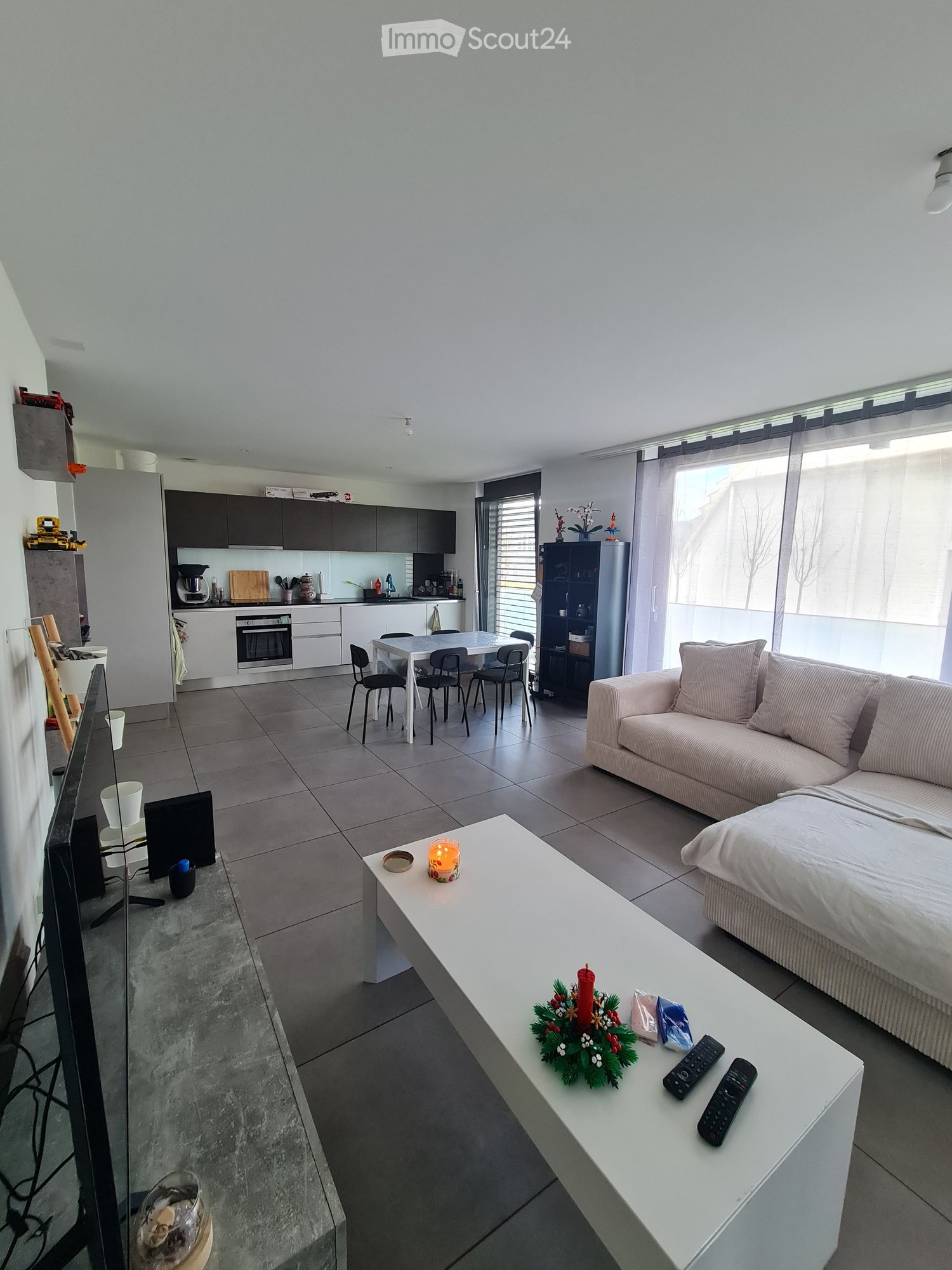 Open plan living room with kitchen, white tiled floor, gray and white sofa, white coffee table, kitchen island with dining table and chairs, black modern appliances, large window