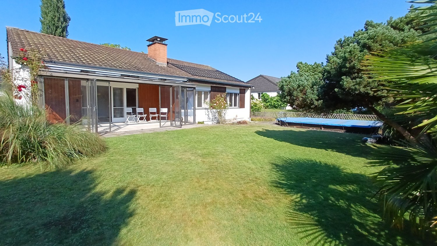 Single family home with lawn, patio with chairs, large trees, a trampoline, roof