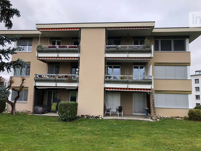 An apartment building with three stories, balconies on all floors, a grassy area, shrubs, and a small garden with a tree in front