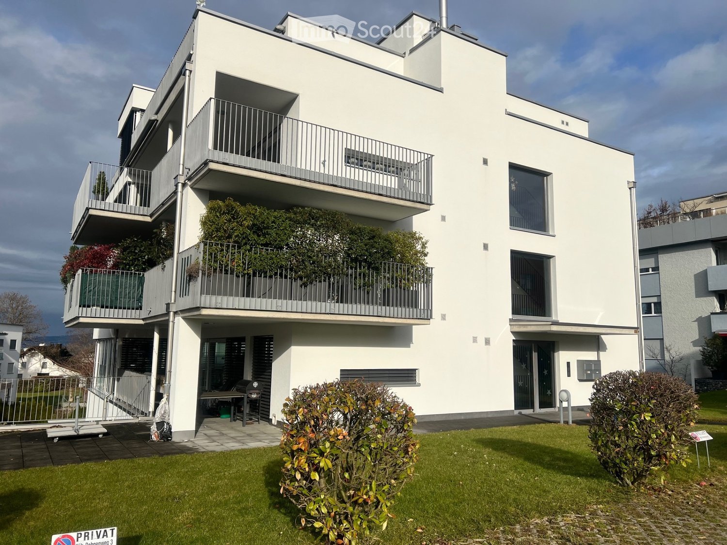 Modern white apartment building with multiple balconies, raised ground floor, and well-maintained green lawn in front.