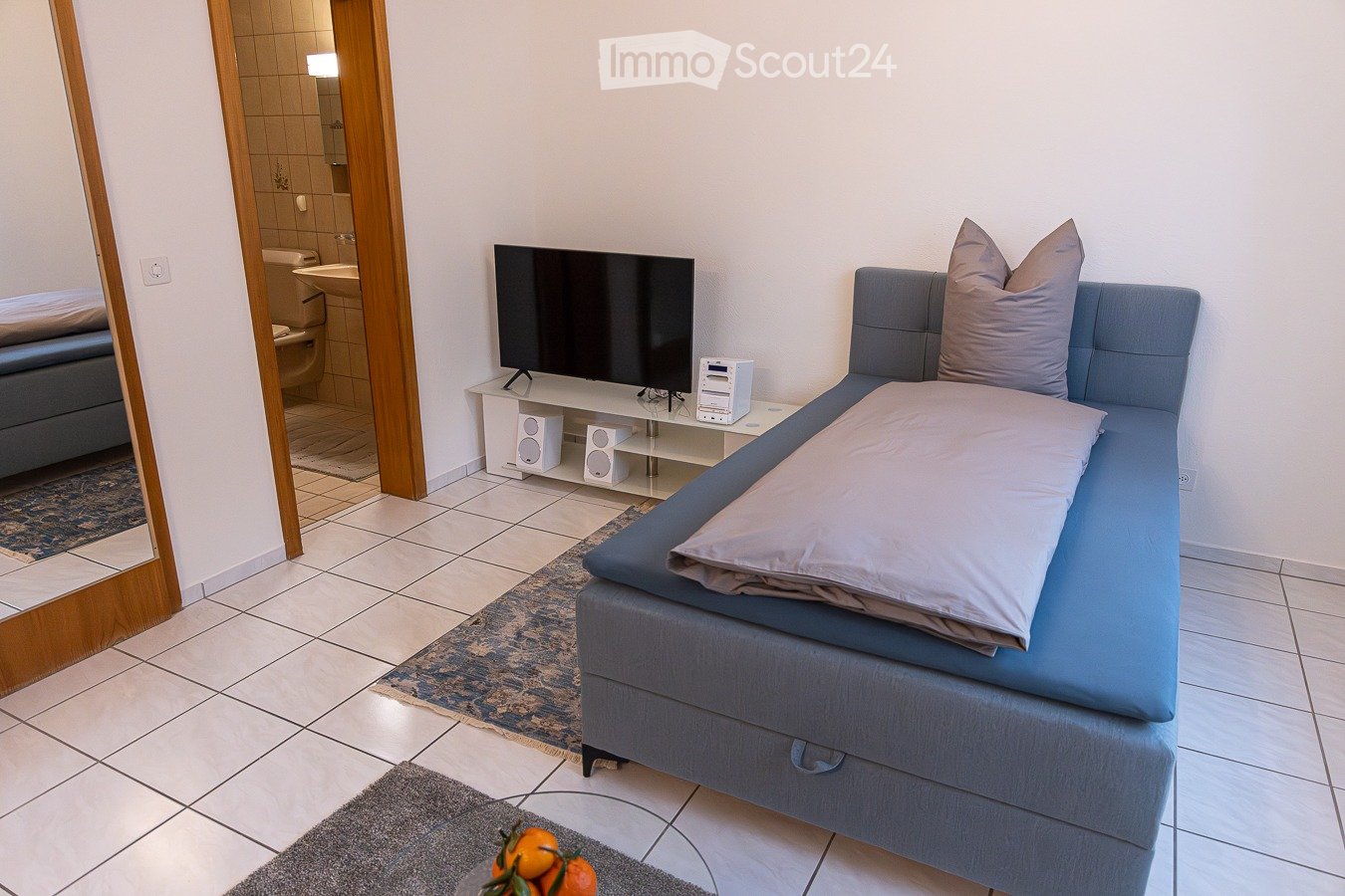 Blue single bed, white tile floor, white walls, modern flat-screen TV, white entertainment center, grey rug, round glass table with oranges, mirror on the wall