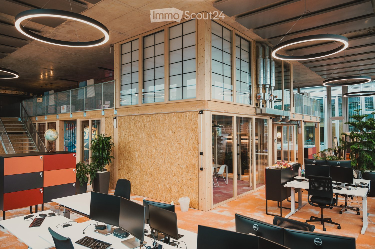 A modern open-plan office with a contemporary design, featuring an array of desks, chairs, and computers. The walls are partially made of wood and glass, providing a bright and spacious feel. Circular lamps hang from the ceiling, and the floor is covered
