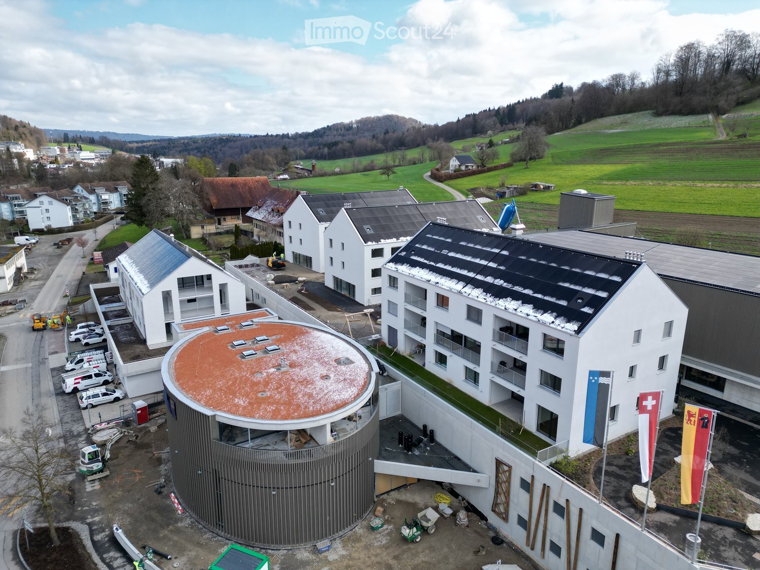 Aerial view of a modern building complex with a circular structure, surrounded by a green field and mountains in the background.
