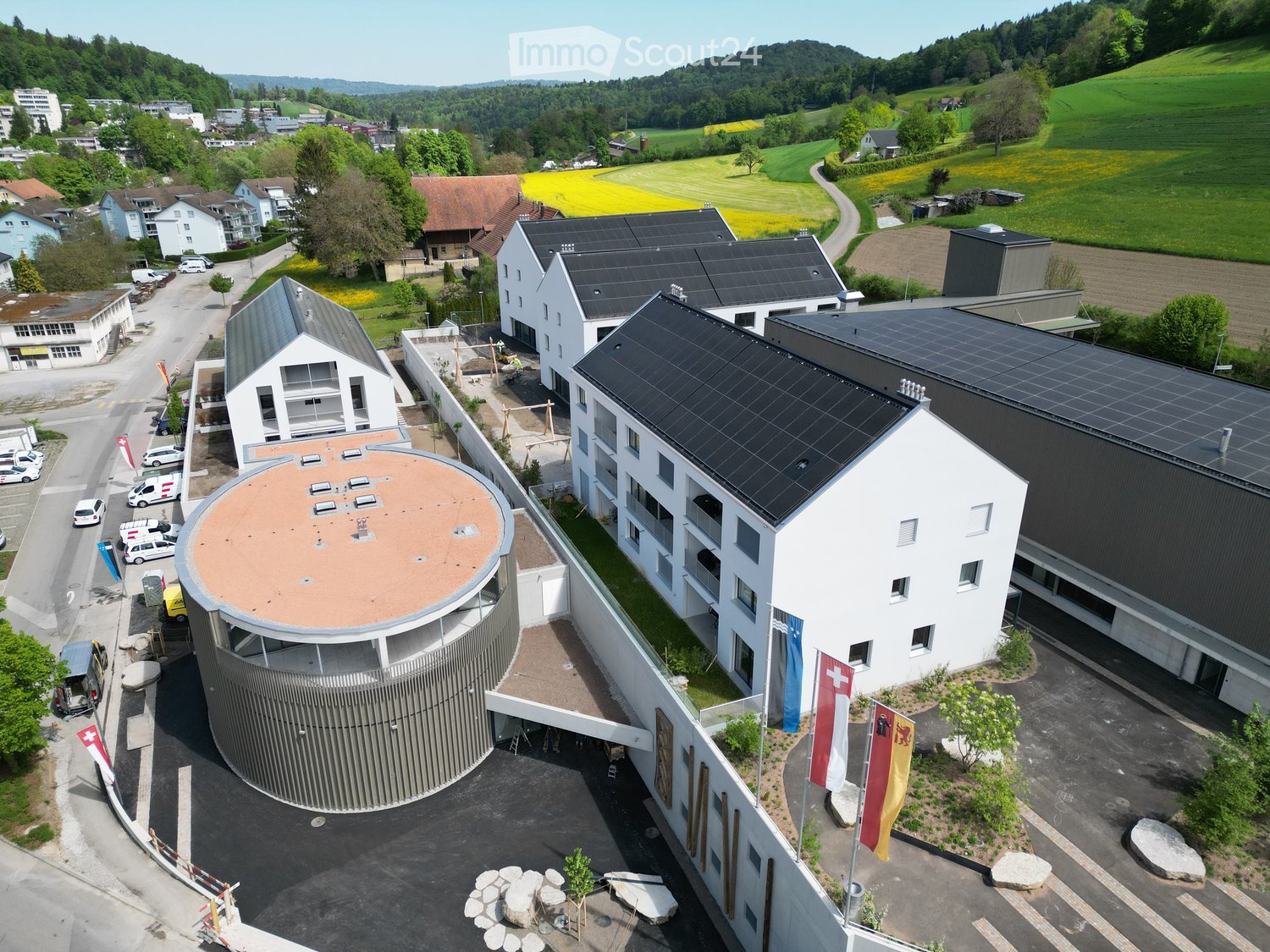 Aerial view of a modern multi-building complex with solar panels on the roofs, surrounded by greenery and a rural landscape.