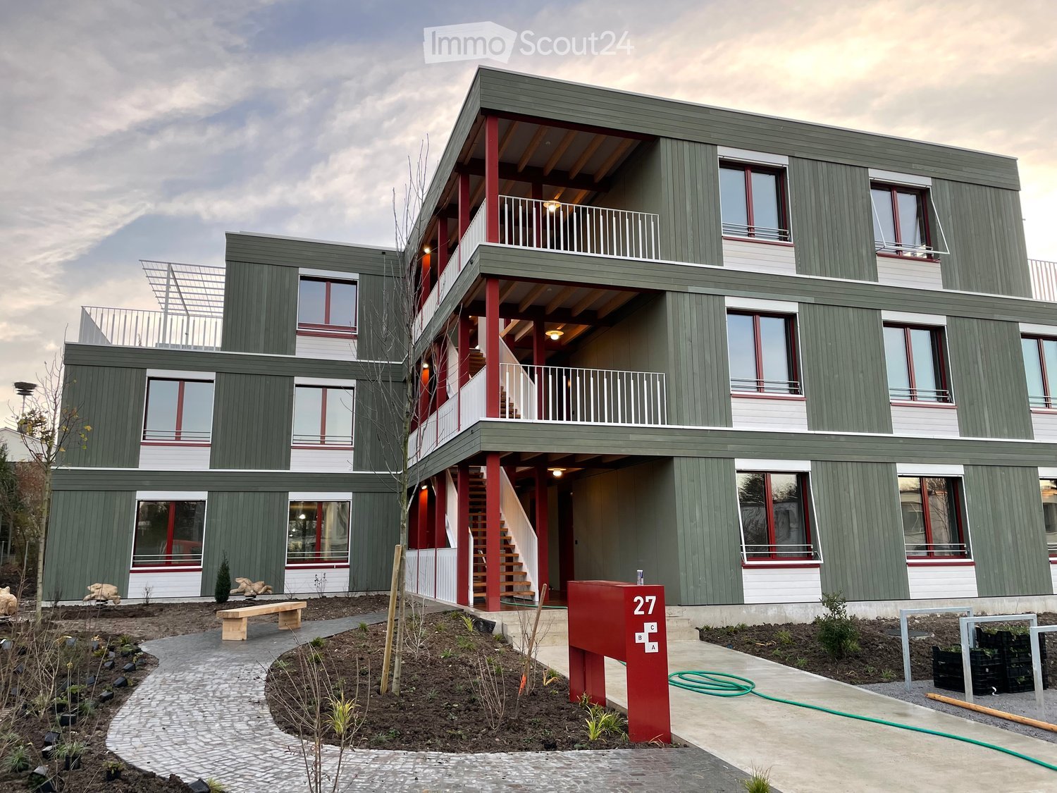 Modern apartment building with green exterior, balconies, windows with red frames, stairs to entrance, small garden in front