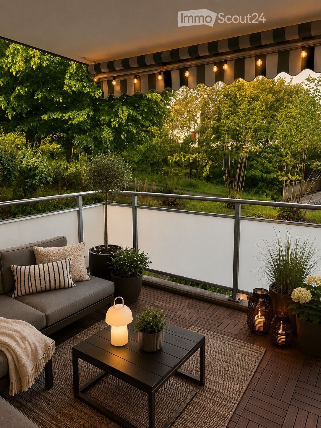 A modern balcony with a grey sofa, coffee table, table lamp, potted plants, and decorative lanterns, surrounded by greenery and a striped awning.