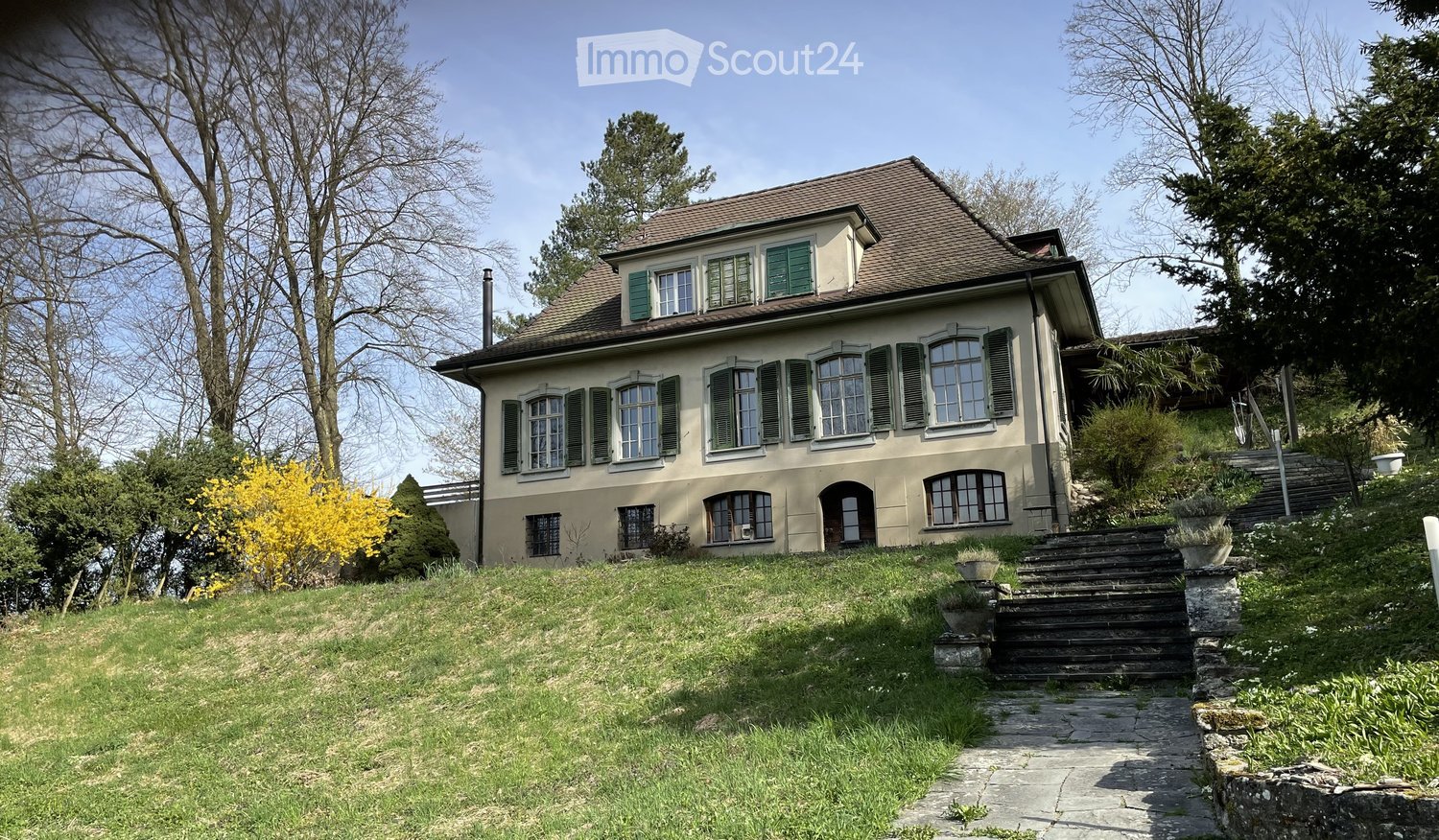 Traditional two-story house, with brown roof, green shutters, raised ground floor, staircase, multiple windows, surrounded by greenery and steps leading up to the entrance