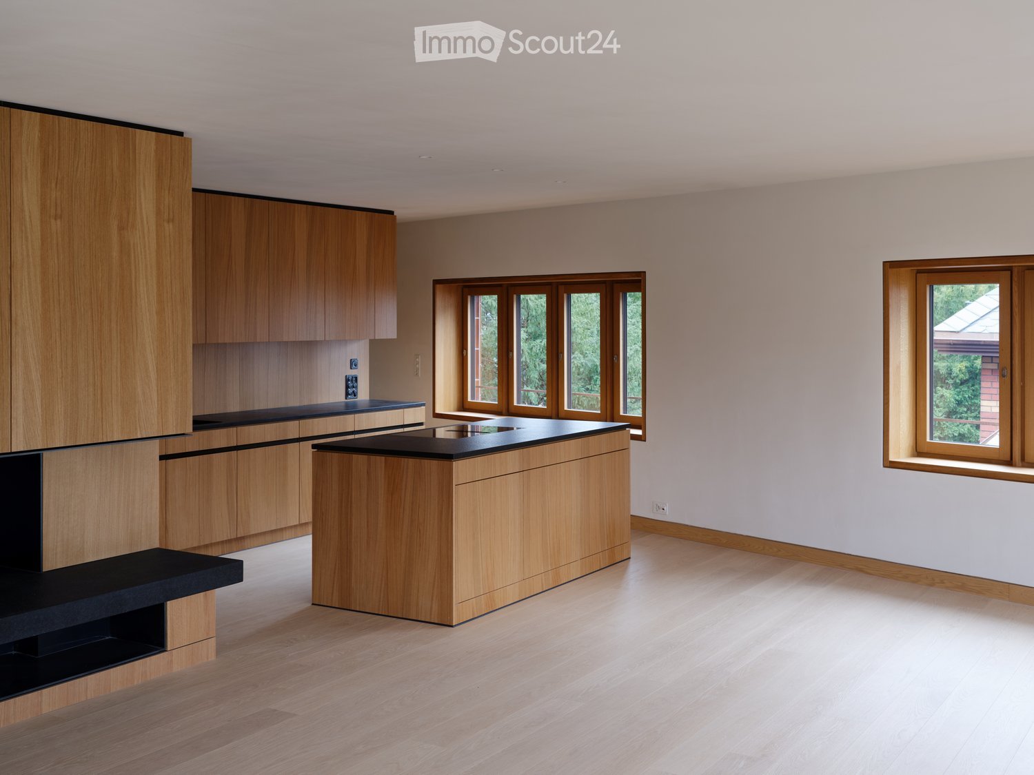 Modern kitchen with light wooden cabinets, black countertops, and large windows allowing natural light.