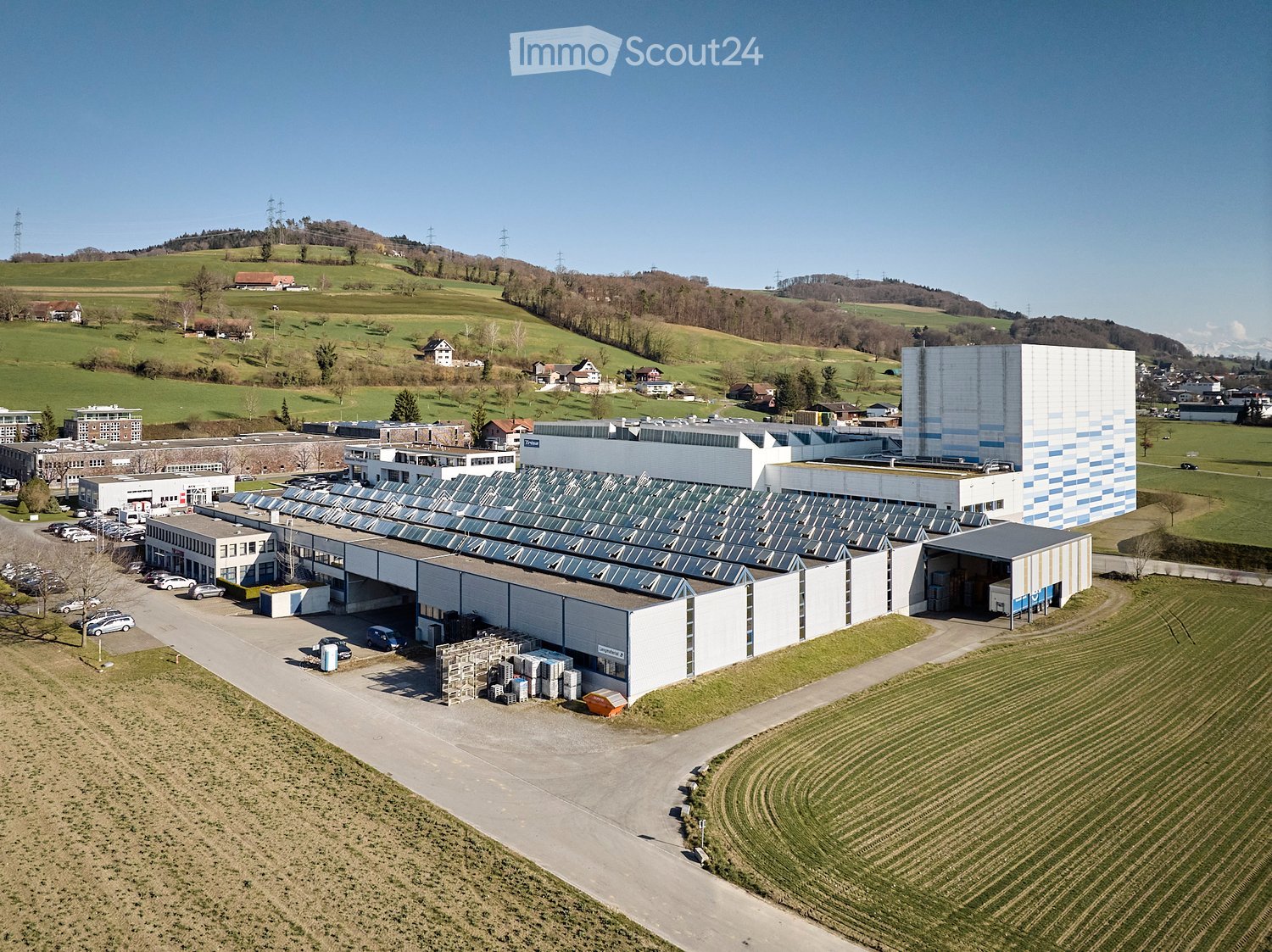 Large industrial building with solar panels on the roof, surrounded by fields and a parking lot