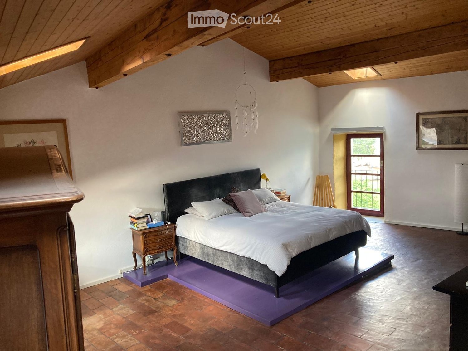 Bedroom with wooden ceiling, white walls, wooden floor, bed with white sheets, nightstand with books, decorative items on the wall, window with a view outside.