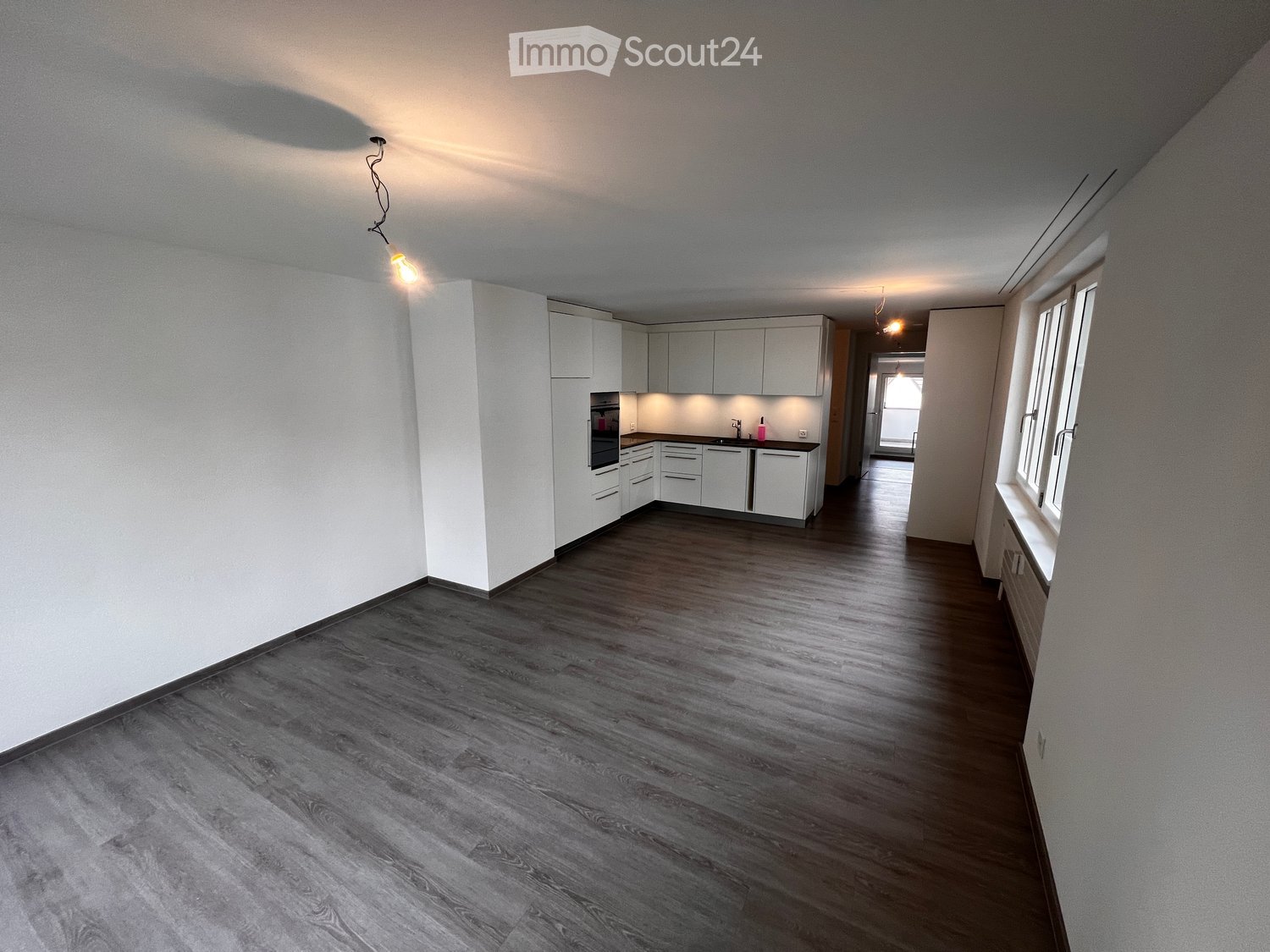 Empty room with white walls, wooden floor, kitchen area with white cabinets, oven, sink, and faucet, and a window with a view.