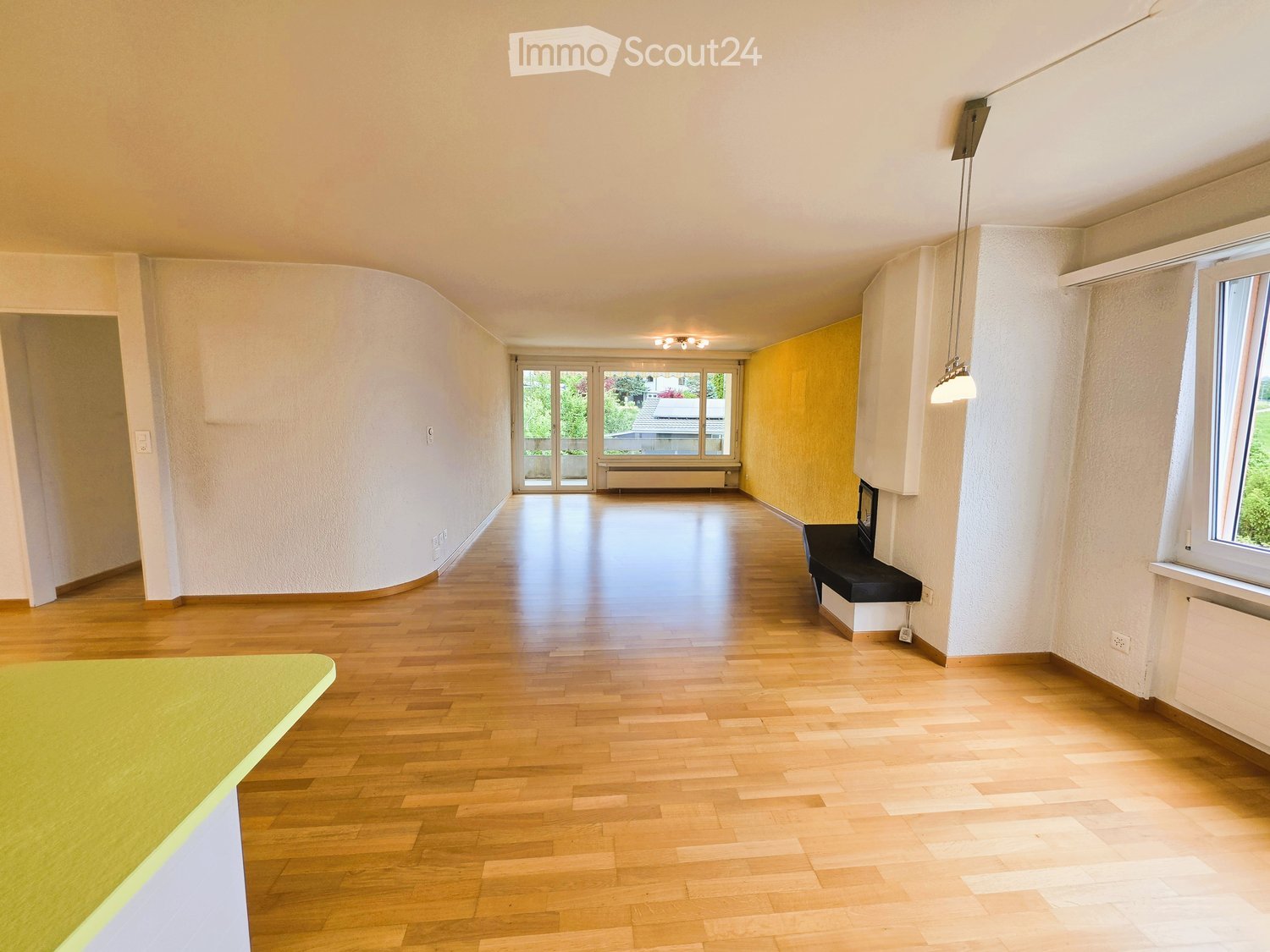 Empty hallway with wooden flooring, white walls, and large windows at the end leading to a balcony area.