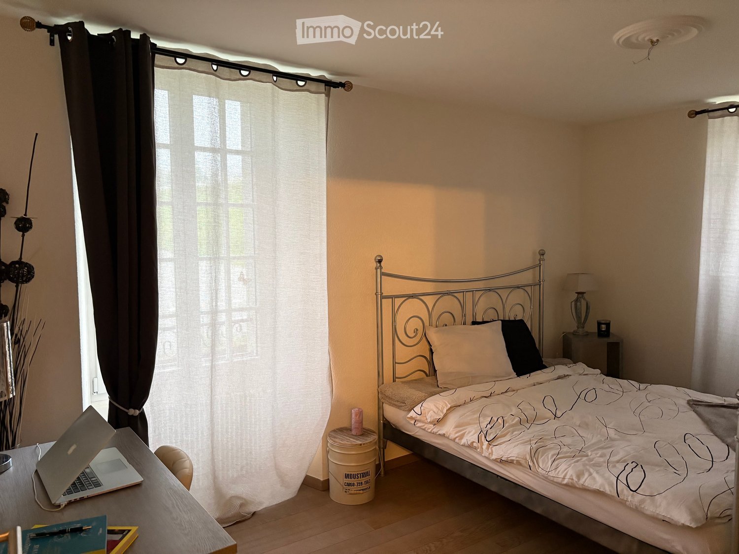 Furnished Room. A bedroom with a bed, desk, laptop, lamp, nightstand, vase, candle, bucket, pillow, and curtains.