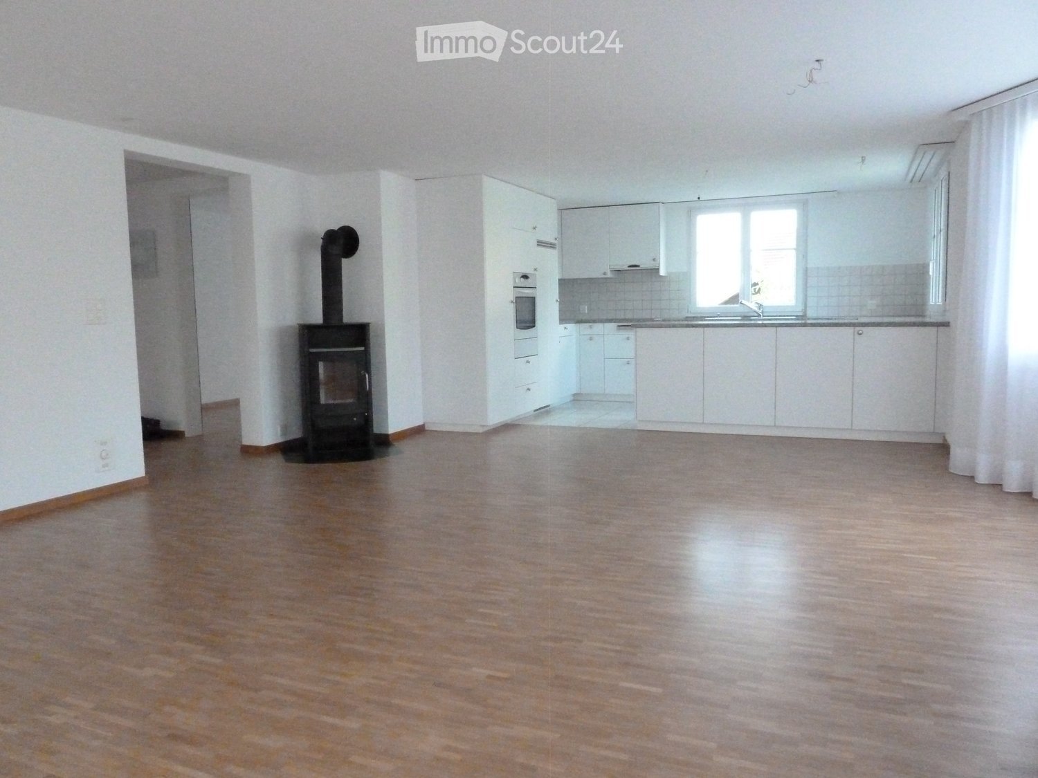 Empty room with wooden flooring, white walls, and a fireplace. The room includes a kitchen area with white cabinets and a sink.