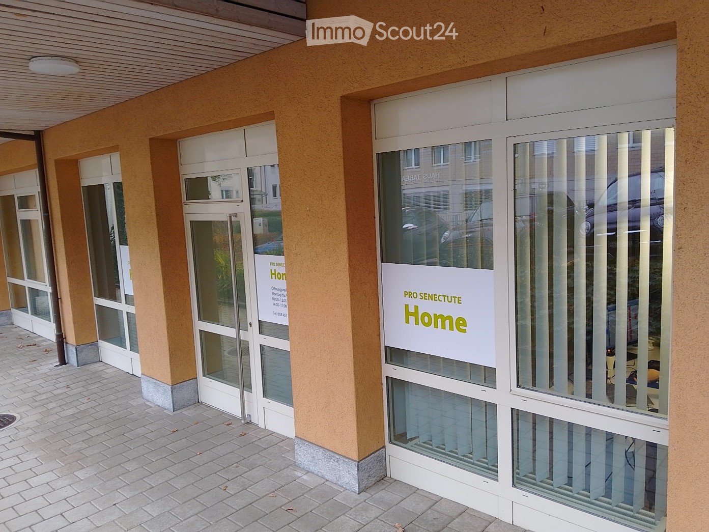 Entrance to a building with glass doors and windows, a sign reading 'PRO SENECUTE Home', and a tiled walkway.