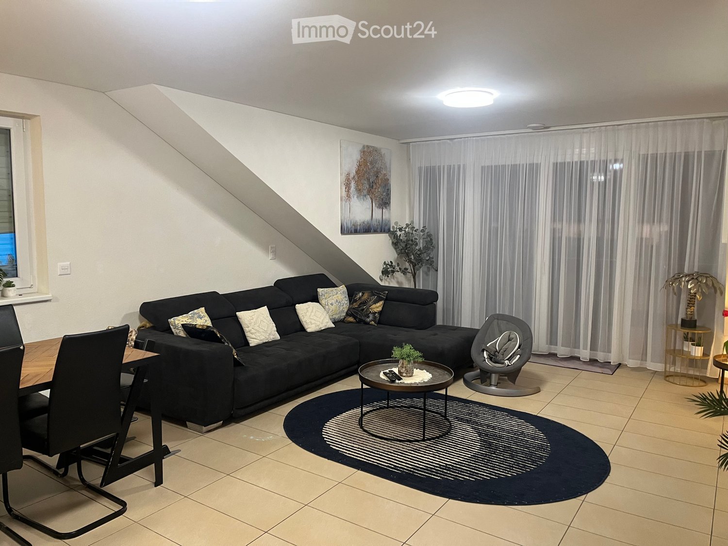 Modern living room with a black sectional sofa, coffee table, baby chair, dining area, and decorative plants.