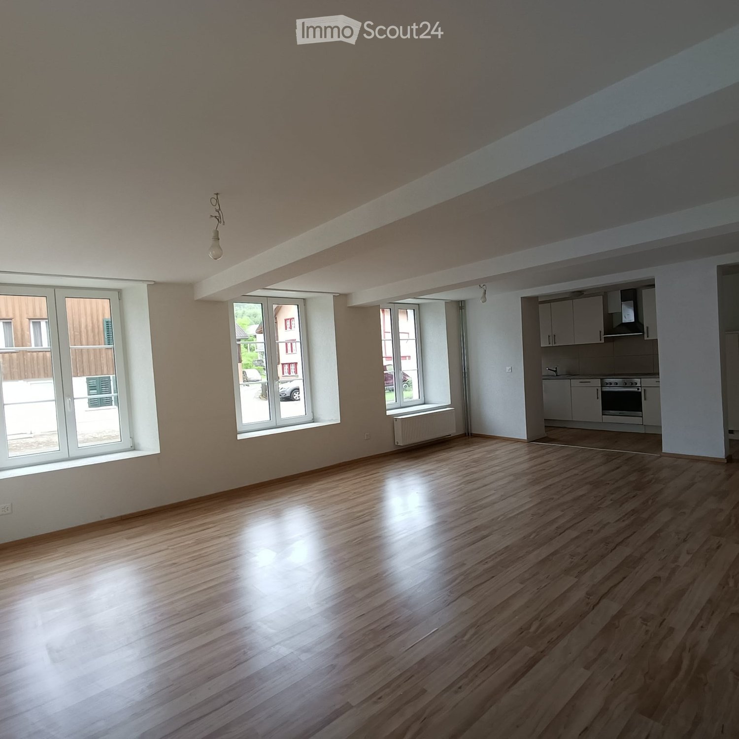 Empty room with wooden flooring, large windows, and a visible kitchen area with cabinets and appliances.