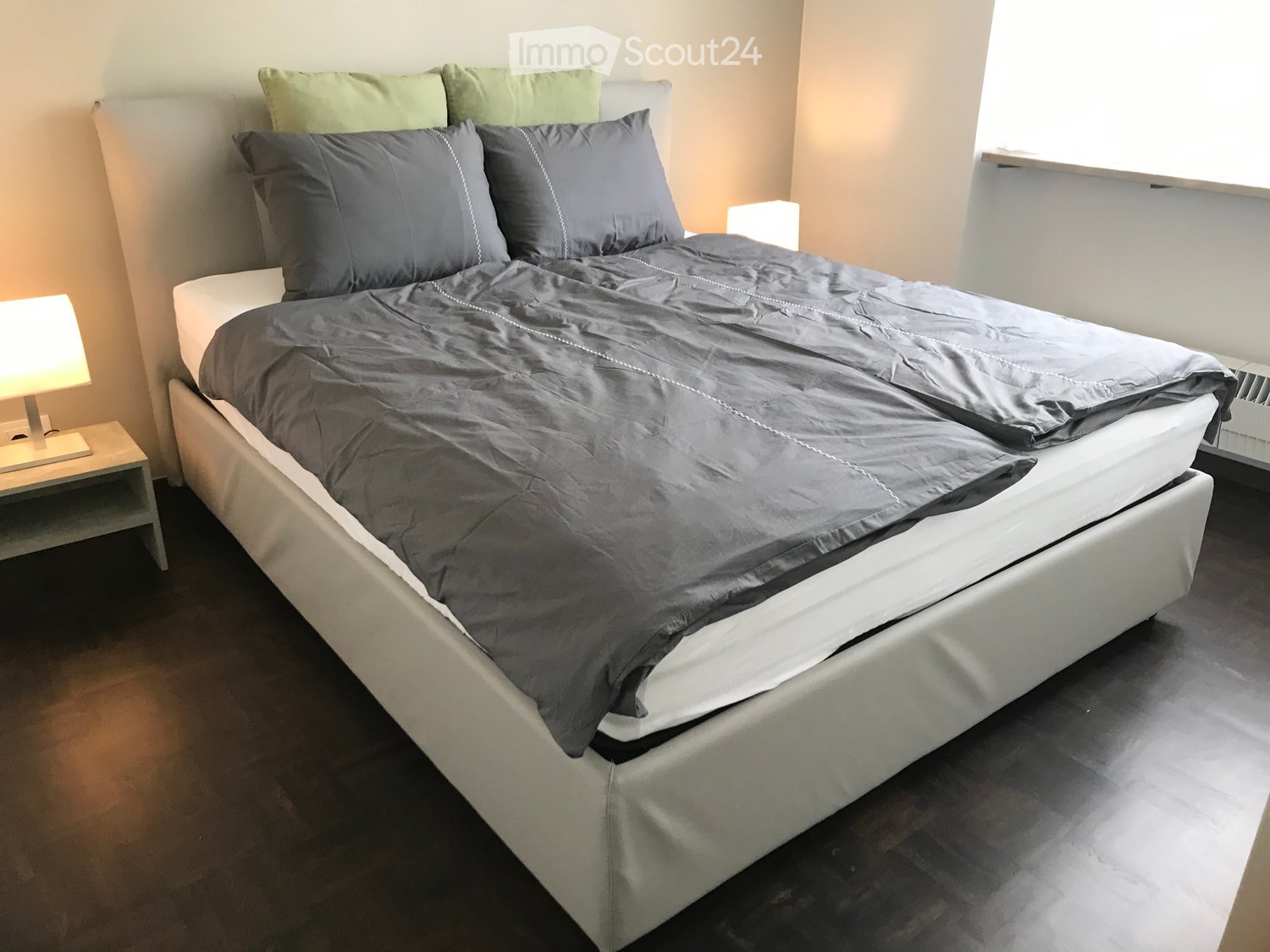 A bed with a white frame, gray bedding, and pillows. Two nightstands with lamps are on either side of the bed. The room has a white wall and a wooden floor.