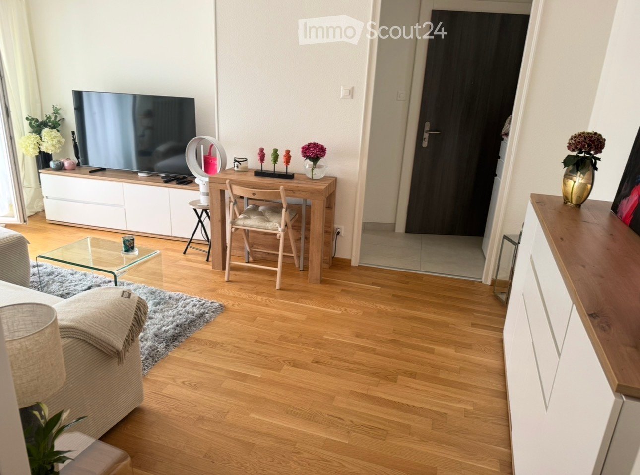living room with wooden floor, white walls, TV, desk, chair, flowers, rug, cabinet, lamp