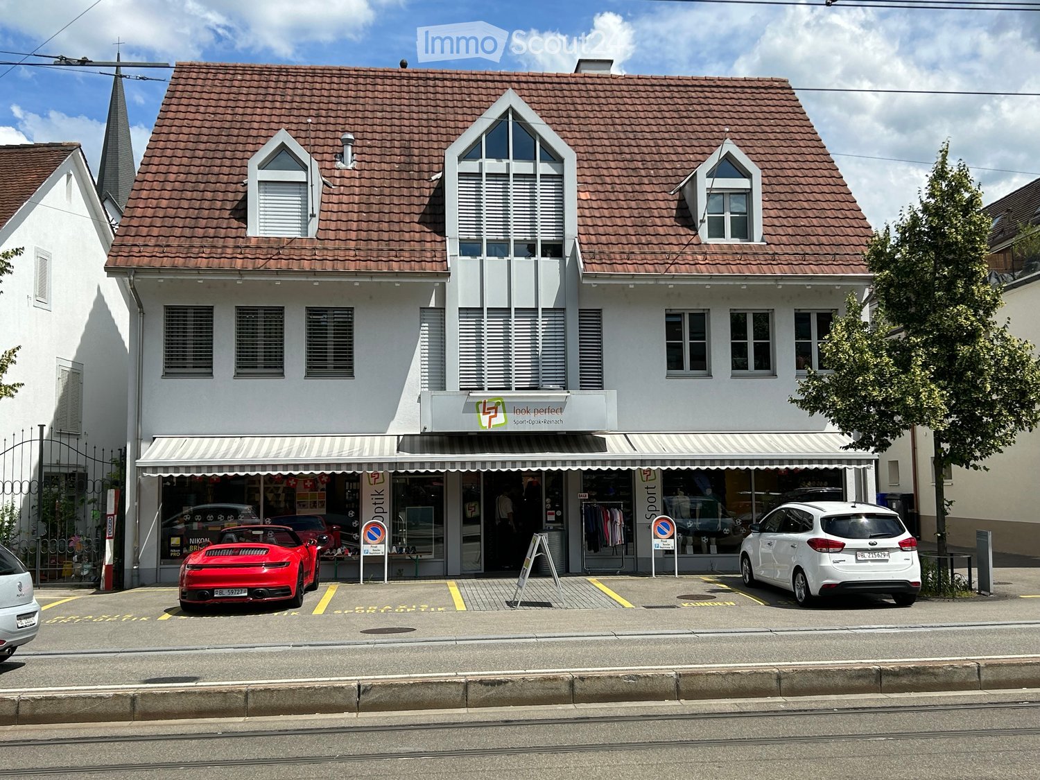 2 story house, red roof, white walls, awning, 2 cars parked in front, shop on the ground floor