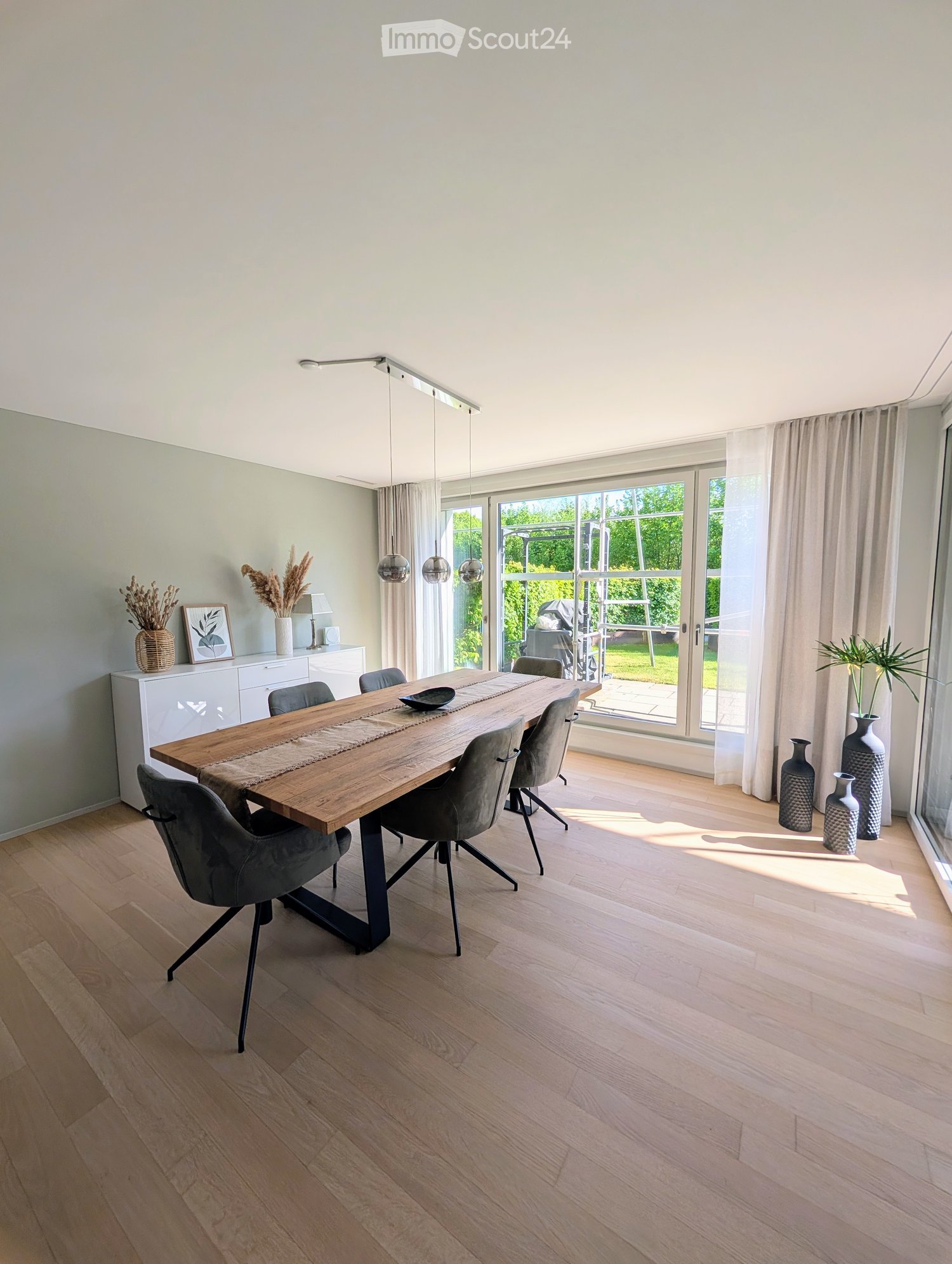 Dining room with wooden table, gray chairs, white cabinets, large windows, vases, and a view of the garden
