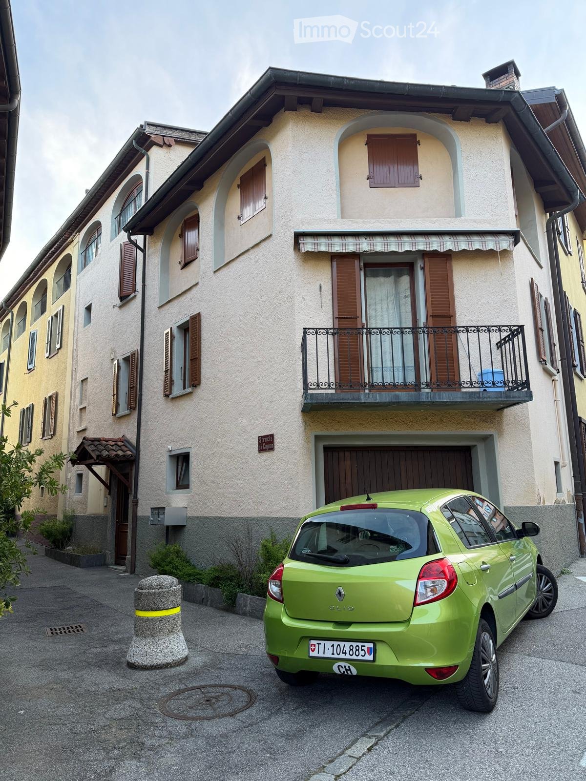 A three-story building with beige walls and brown shutters. A green car is parked in front of the building.