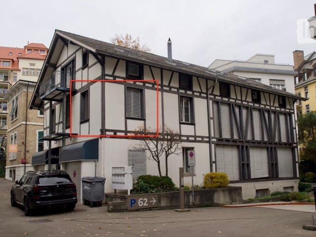 A three-story building with a red square highlighting a specific apartment on the second floor. The building has a mix of white and brown exterior walls, with a balcony on the left side. A black car is parked in front of the building, and a trash bin is p