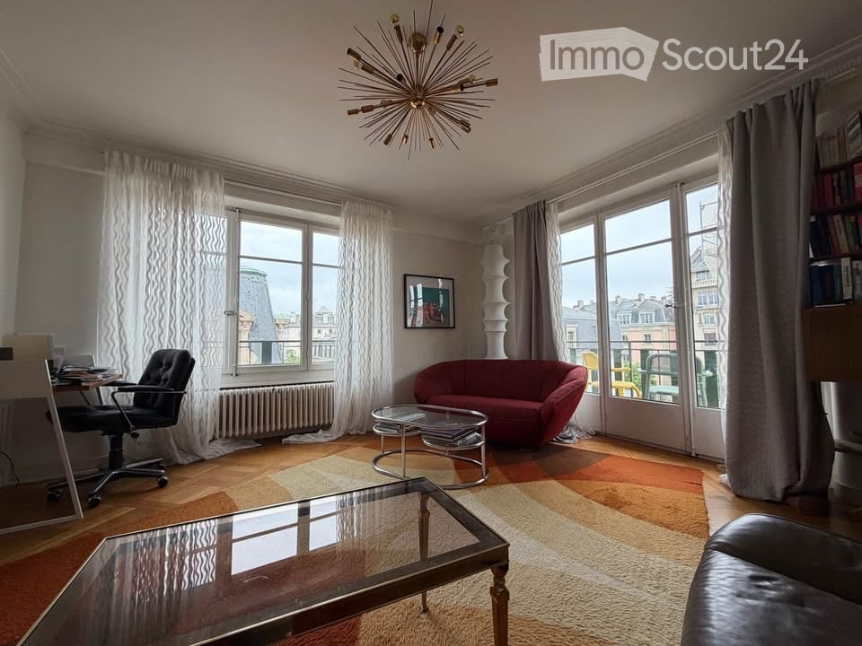 Spacious room with a desk, office chair, red couch, coffee table, and a bookshelf. Large windows with white curtains.