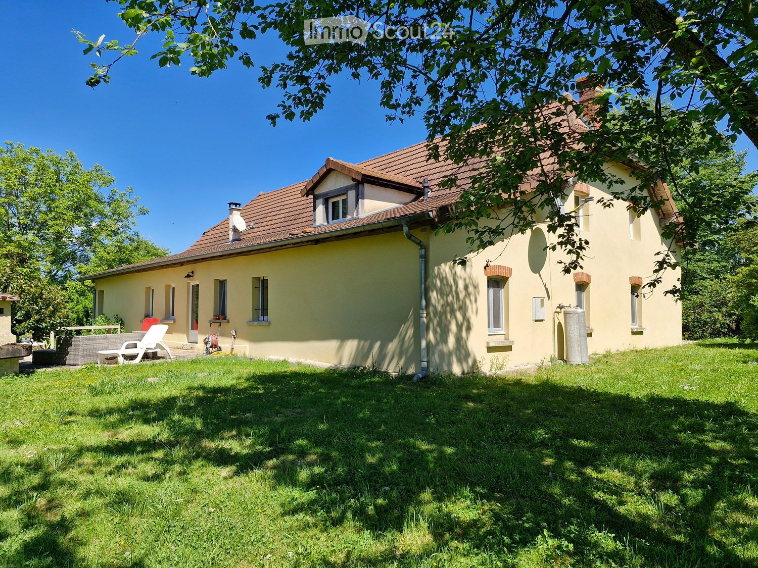 House. Single-story house with yellow walls, red-tiled roof, multiple windows, and a small garden area with outdoor furniture.