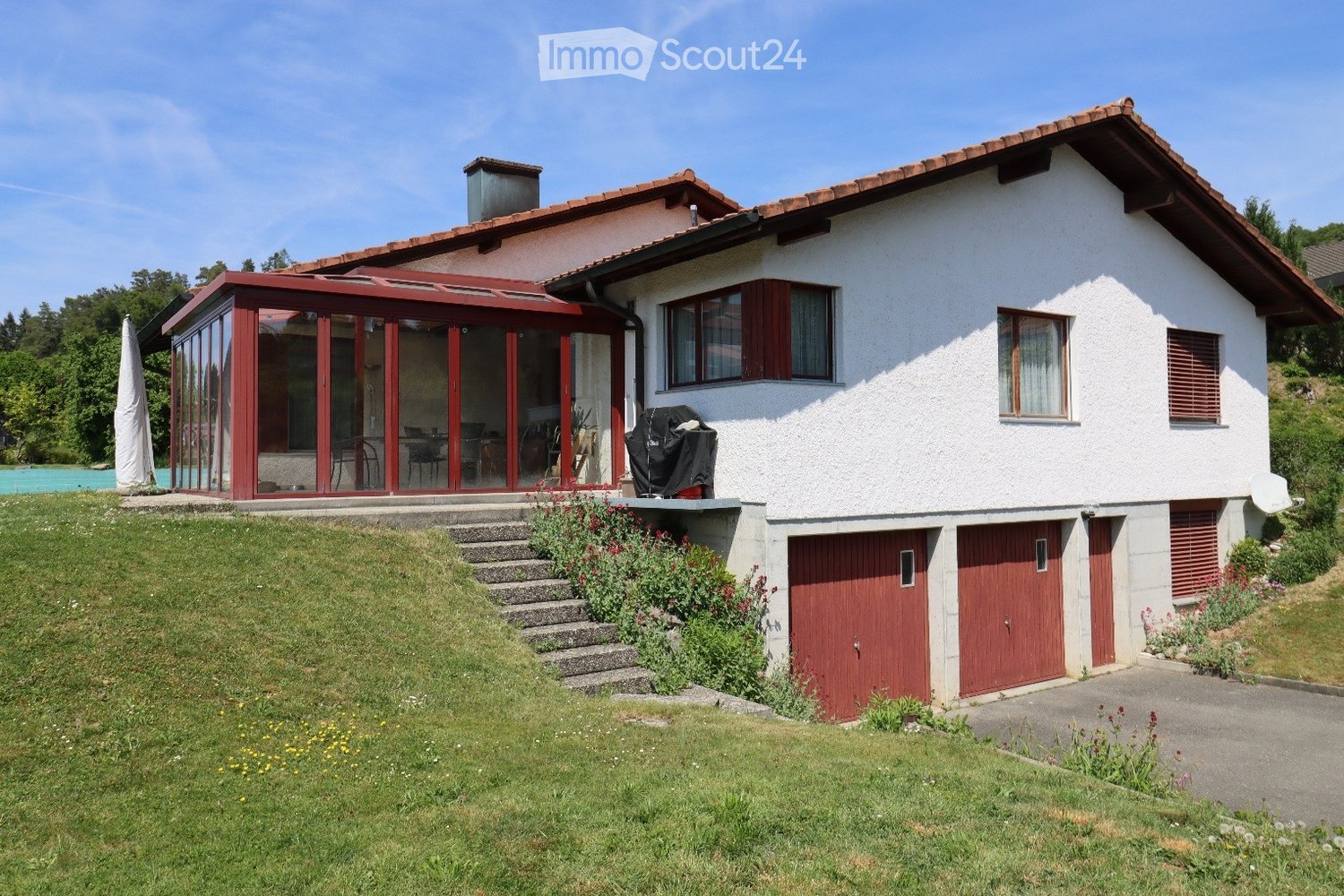 Two-story house with white exterior, red garage doors, and a covered patio area with glass walls. The house features a sloped roof, multiple windows, and a well-maintained garden with steps leading up to the entrance.