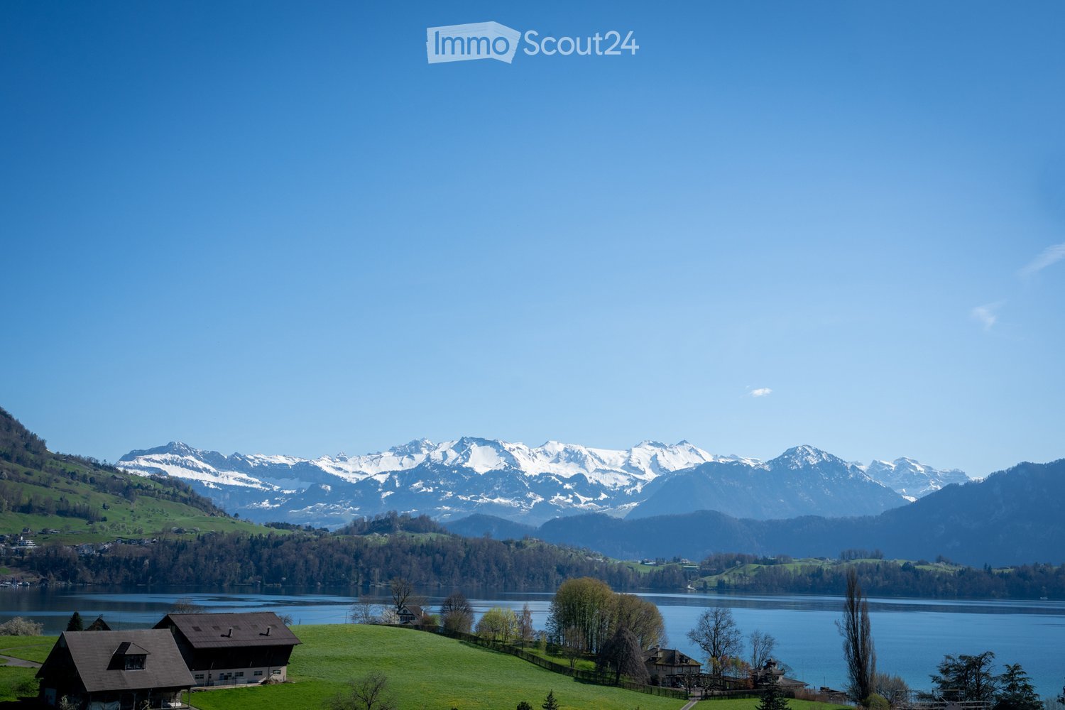 Garden / Outdoor Area. A serene lakeside view with snow-capped mountains in the background, surrounded by green hills and scattered houses.