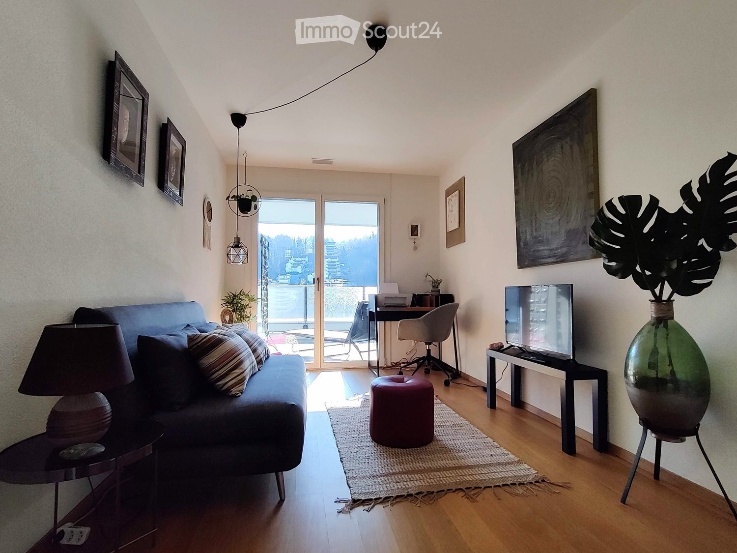 Gray sofa, two side tables with lamps, rug, red foot stool, black TV stand, flat-screen TV, glass door with balcony, black office chair, desk, green plant