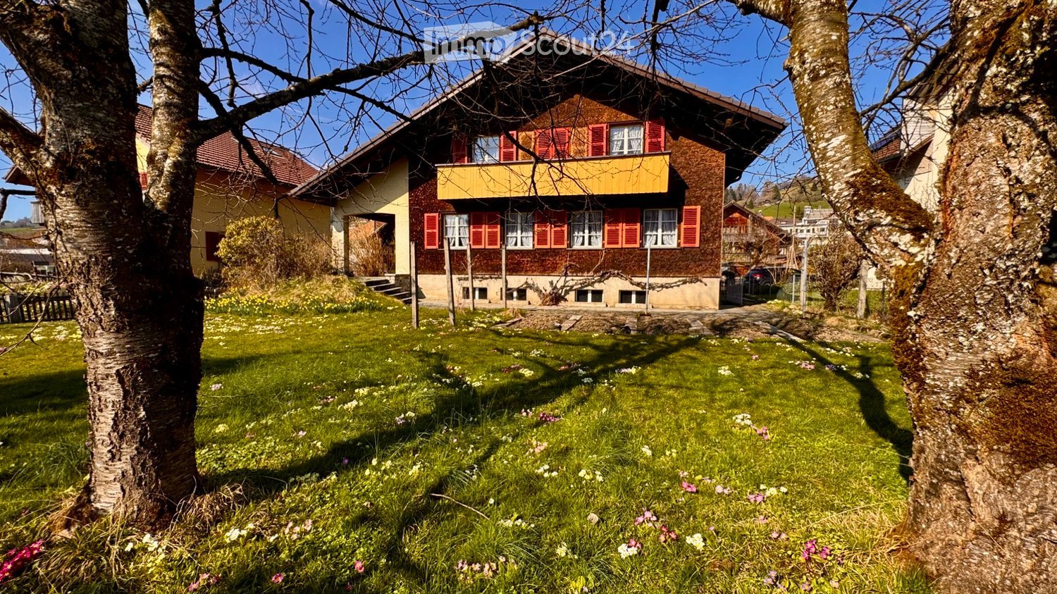 House. A two-story wooden house with red shutters and a yellow balcony, surrounded by a well-maintained garden with grass, flowers, and trees.
