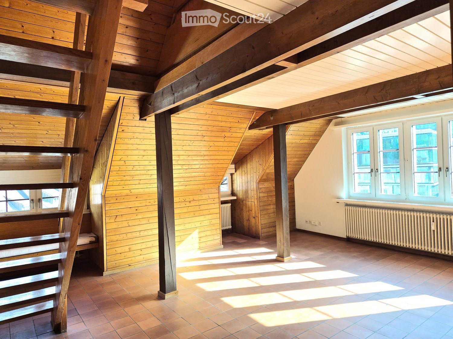 An unfurnished attic room with wooden walls, exposed wooden beams, a wooden staircase, and large windows allowing natural light to fill the space.