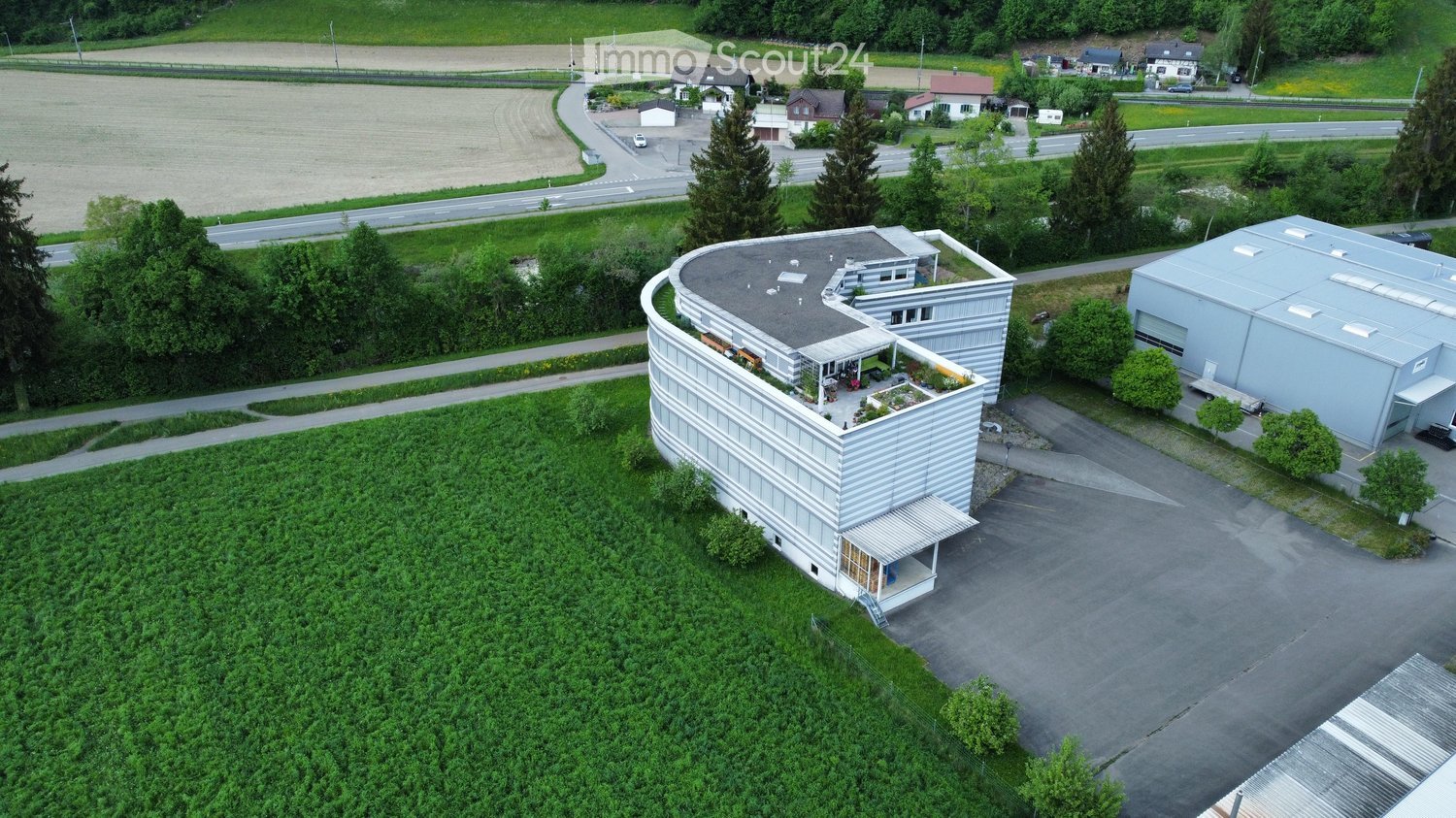 Building. A modern, curved building with a flat roof and a small garden area on the roof, surrounded by greenery and a parking lot.