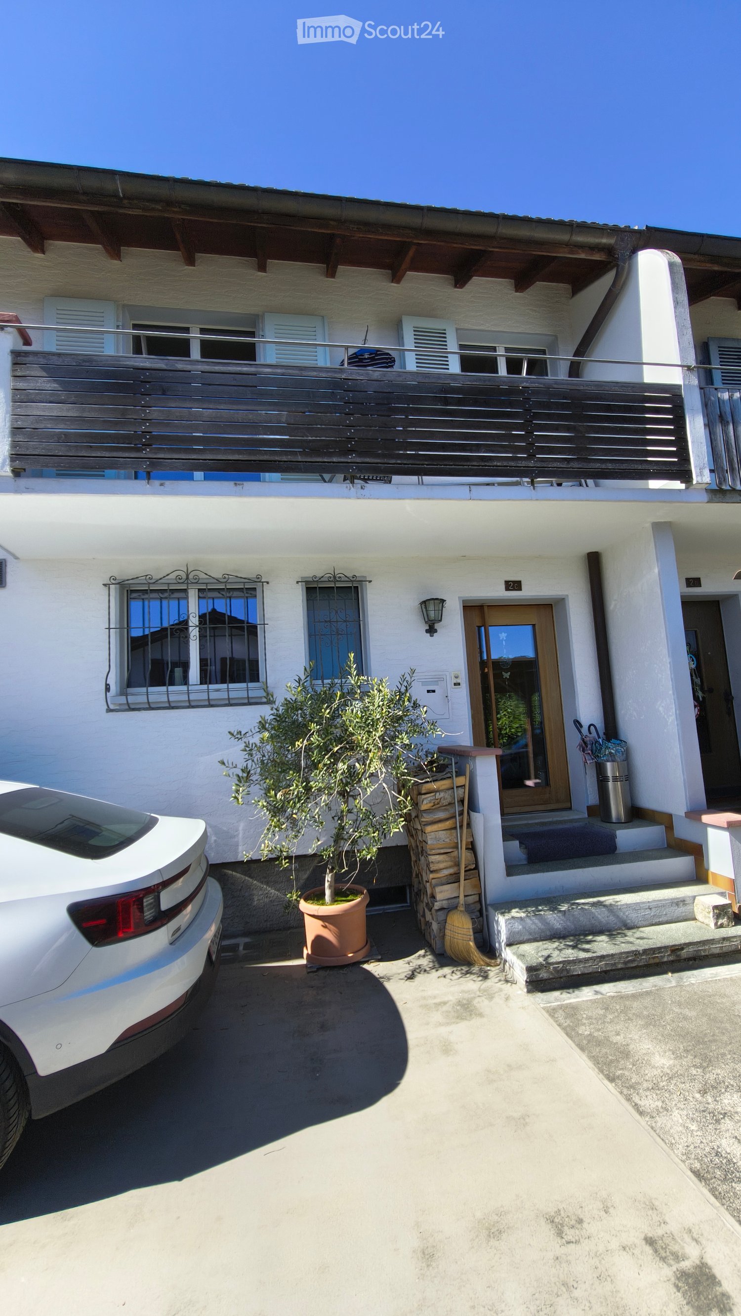 House. Two-story house with white exterior, wooden balcony, and small tree in a pot near the entrance.