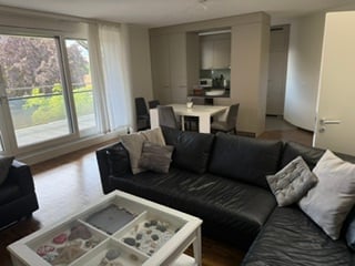Furnished Room. Modern, open-plan living and dining area with large windows offering a view, a black sectional sofa, a white coffee table, a dining table with chairs, and contemporary kitchen cabinets.