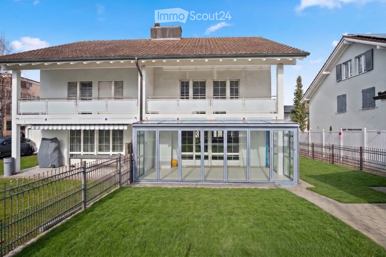 House. Two-story house with a tiled roof, white exterior, multiple windows, and a covered patio area with glass walls. The property features a well-maintained lawn, a metal fence, and a covered outdoor seating area.