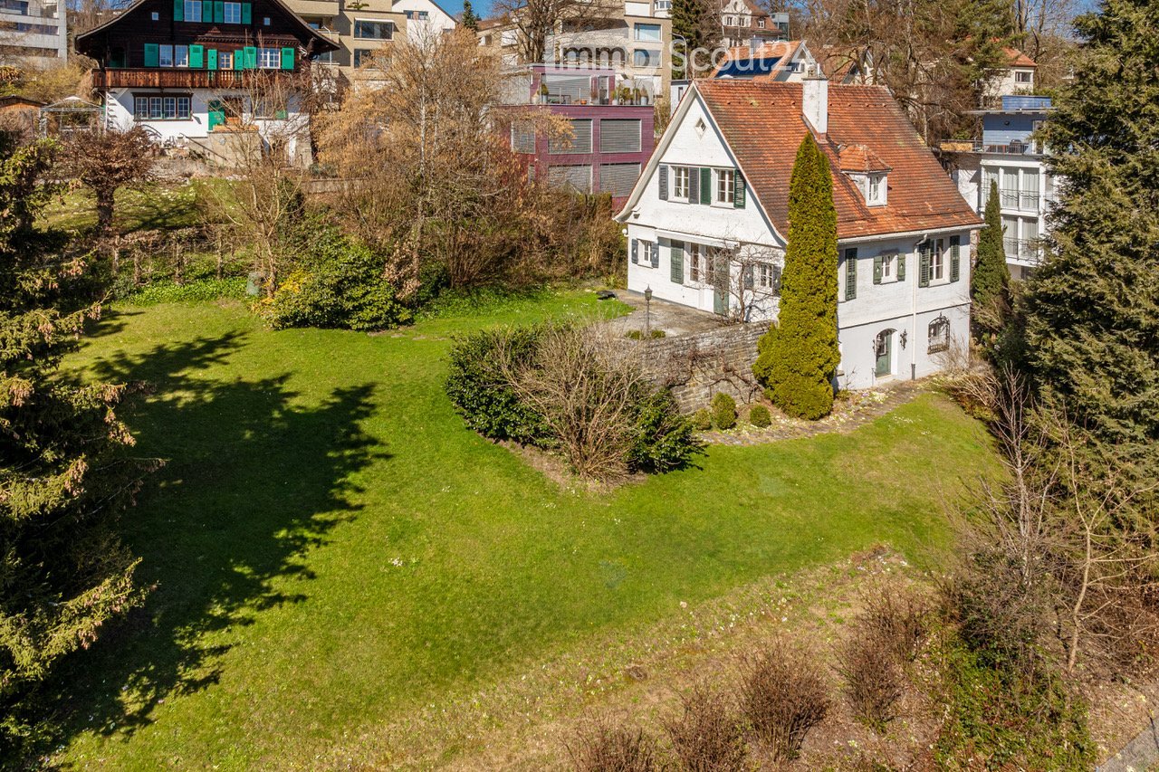 Garden / Outdoor Area. Aerial view of a well-maintained garden with a large green lawn, surrounded by trees and bushes. Several houses with distinct architectural styles are visible in the background, including a white house with green shutters and a red-