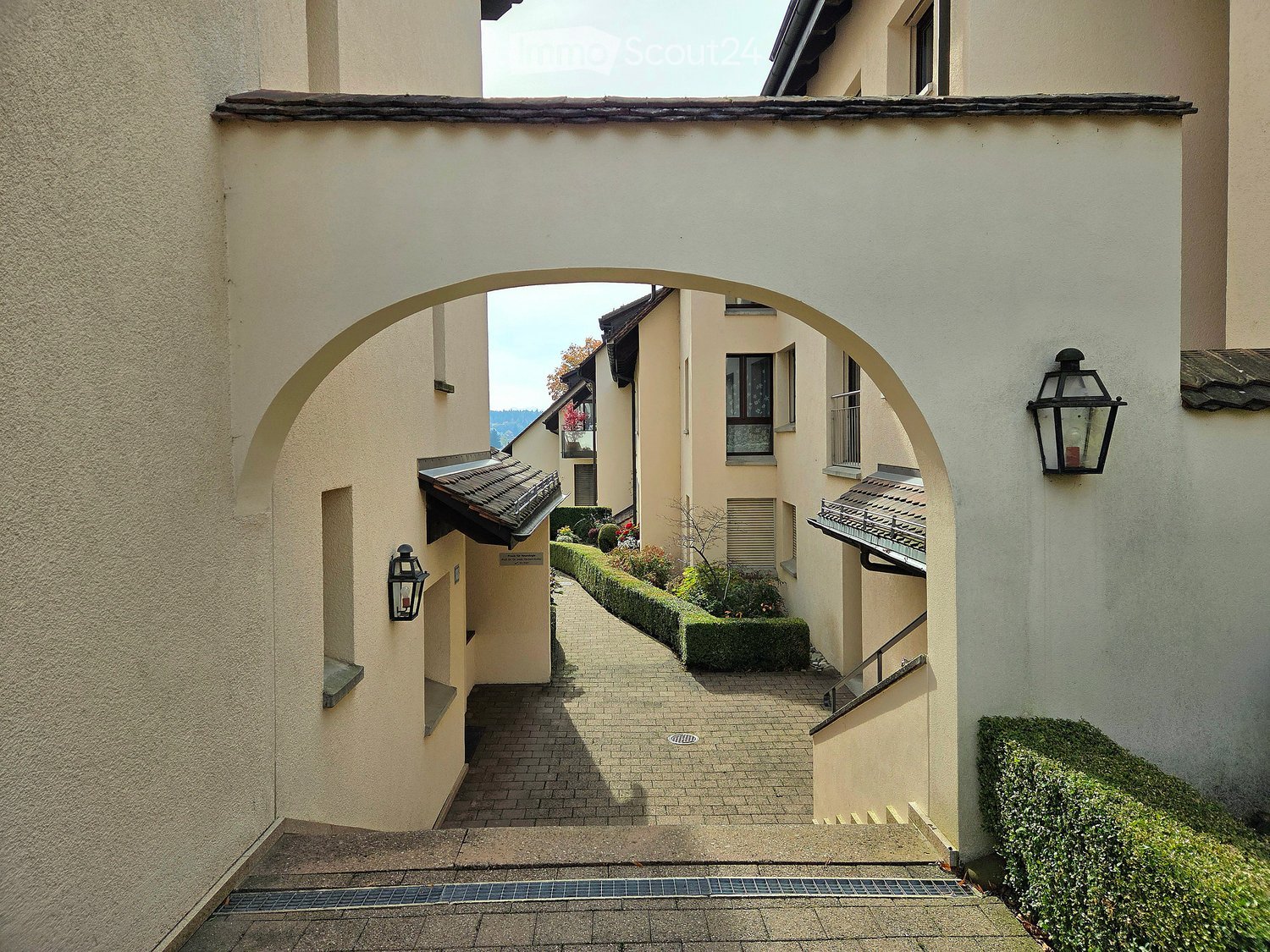 A narrow, arched corridor with beige walls, brick pavement, and small garden beds on either side. Lantern-style wall lights are mounted on the walls.